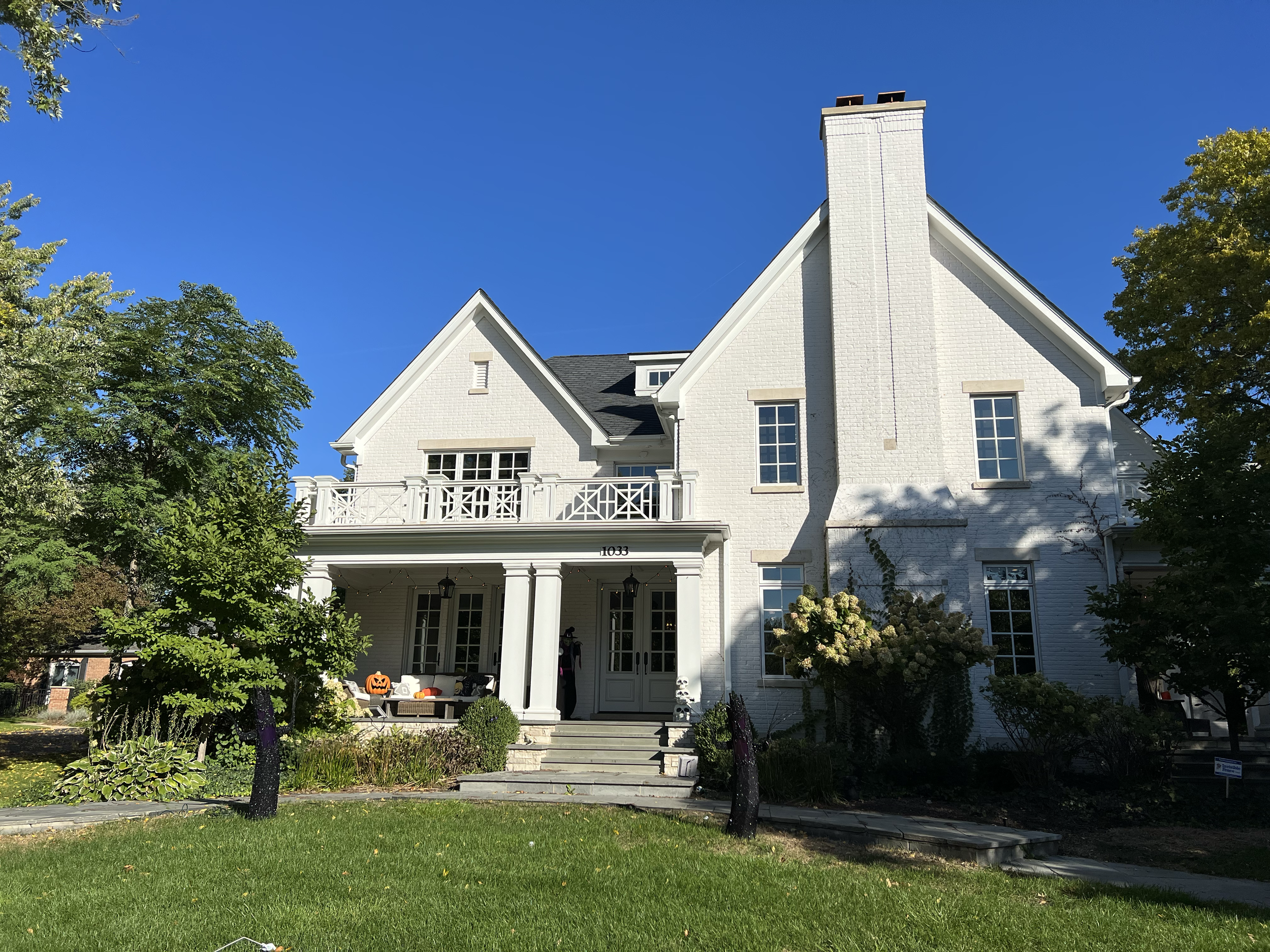White two-story house with a front porch, several windows, a balcony, a staircase, and Halloween decorations including a pumpkin, on a sunny day with a blue sky, surrounded by green trees and grass.