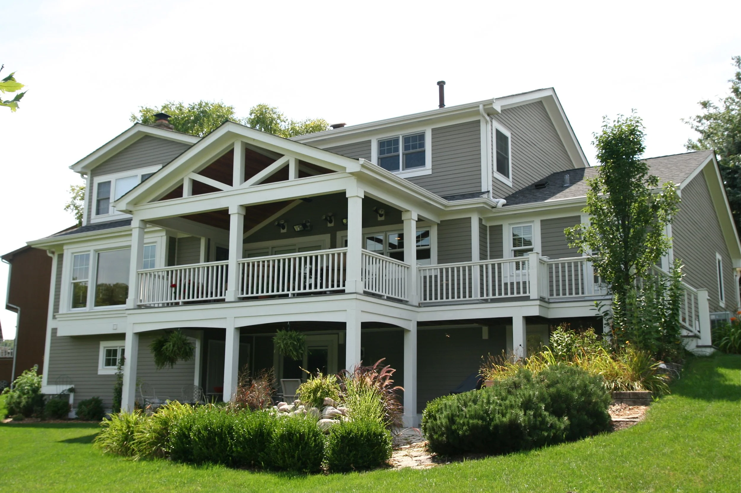 Large gray house with multiple levels and white railings, surrounded by green lawn and landscaped bushes and trees.