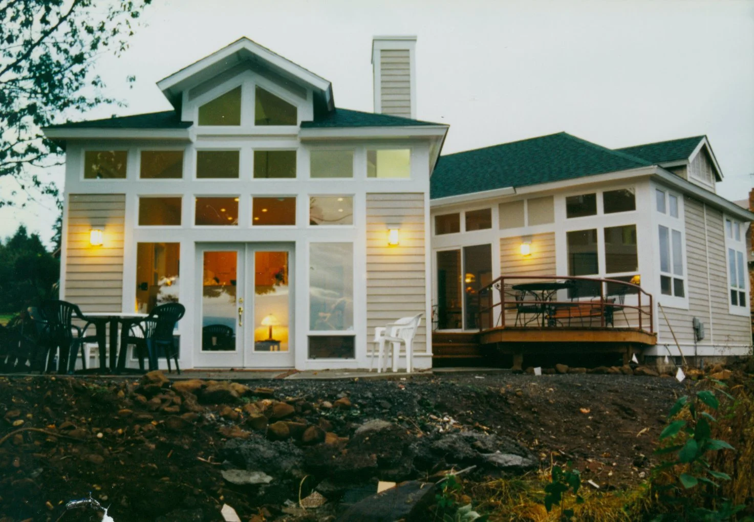 A two-story house with large windows, outdoor seating areas, and a small deck, illuminated by exterior lights, with a rocky and partially landscaped yard in the foreground.