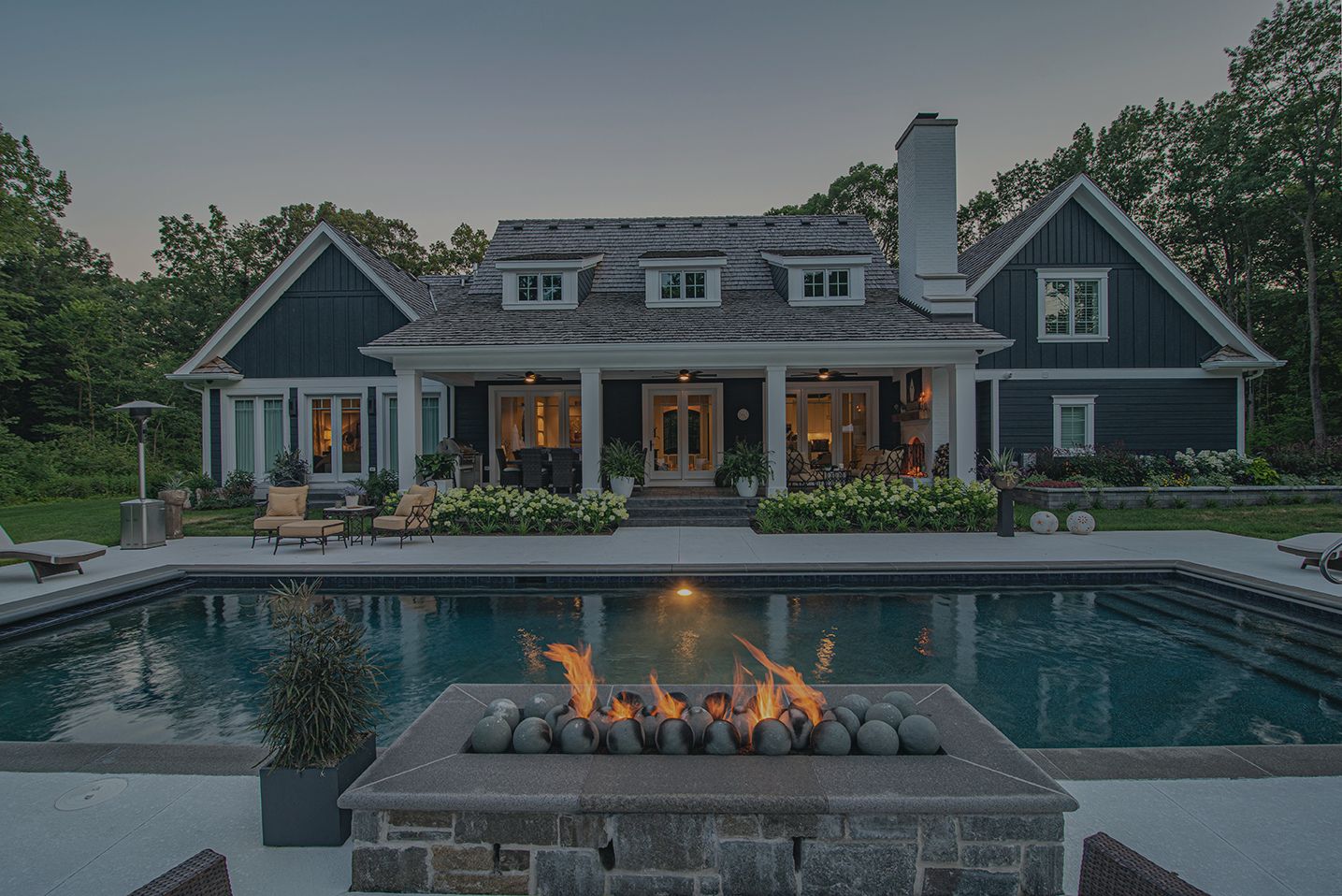A large modern house with a dark exterior, multiple windows, and a chimney. In front, there's a swimming pool with lounge chairs, a fire feature with flames, and lush landscaping with plants and flowers.
