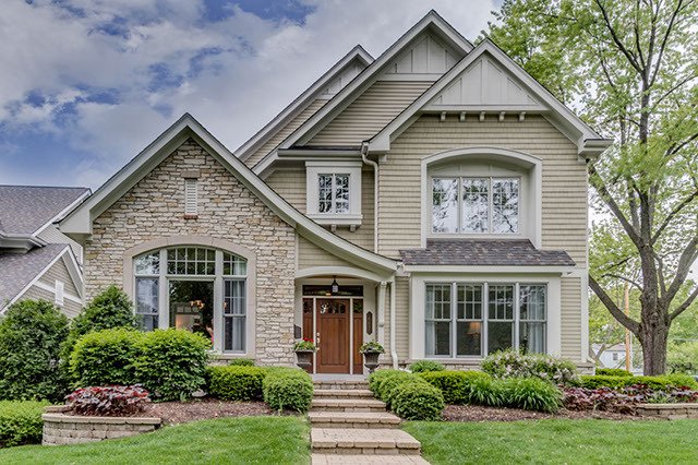 Front view of a two-story suburban house with beige and stone exterior, large windows, a wooden front door, and a well-maintained lawn with bushes and trees.