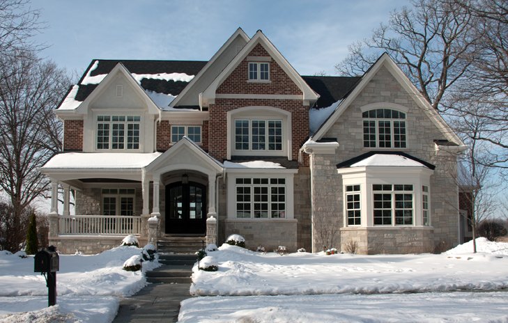Large snowy house with a stone and brick facade, multiple gabled roofs, and a front porch, surrounded by snow-covered lawn and leafless trees.