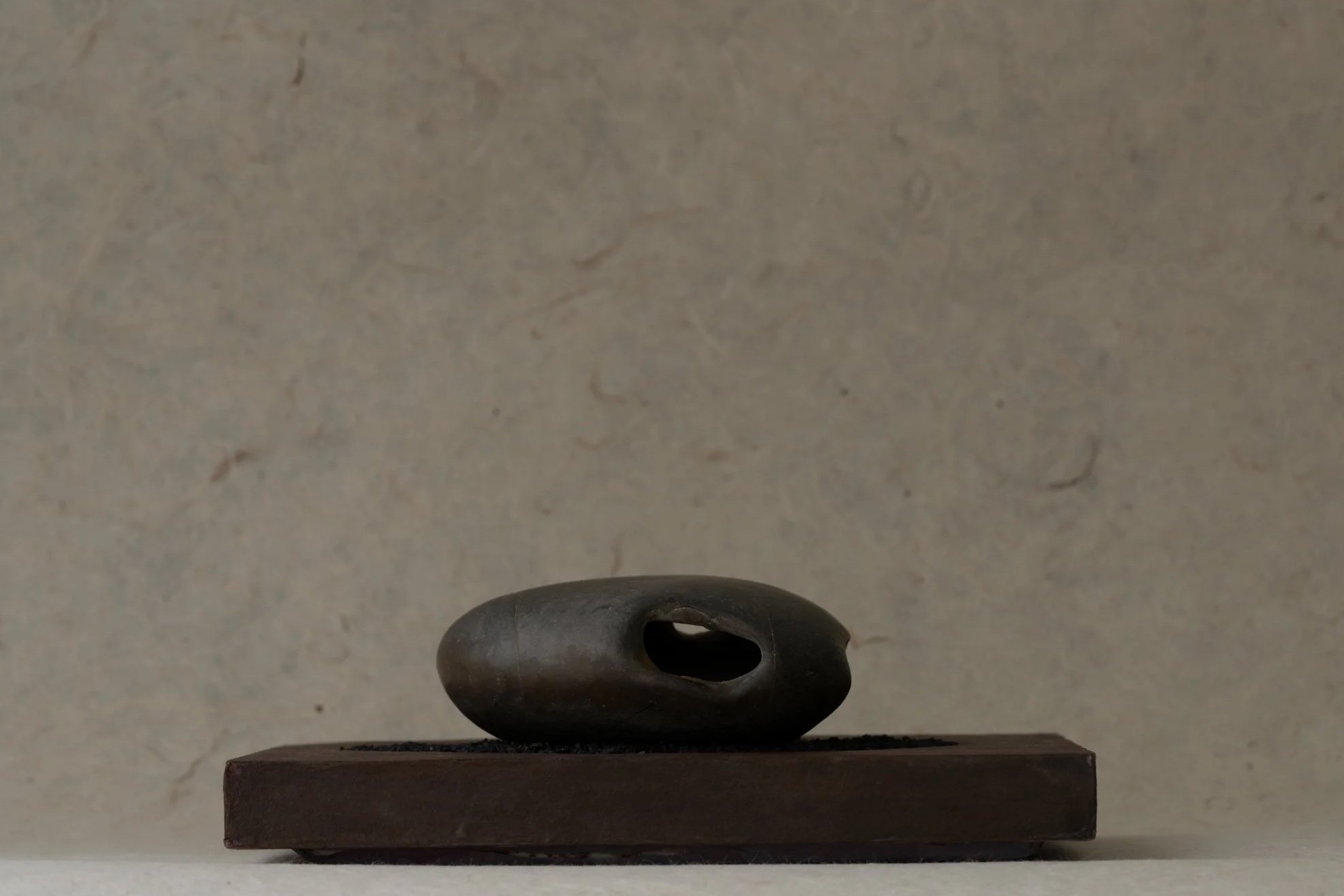 A small black sculpture with an oval shape and hollow center, positioned on a rectangular base, against a light-colored textured background.