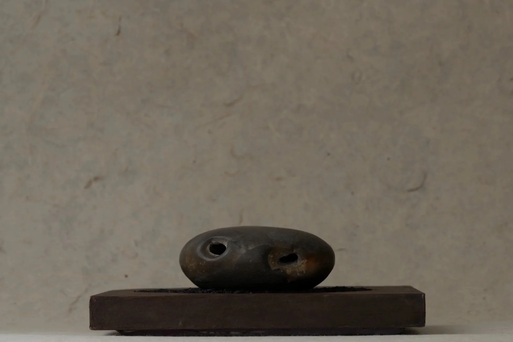 A small, dark-colored sculpture of an oval shape with multiple holes, placed on a rectangular brown base, against a plain light-colored background.