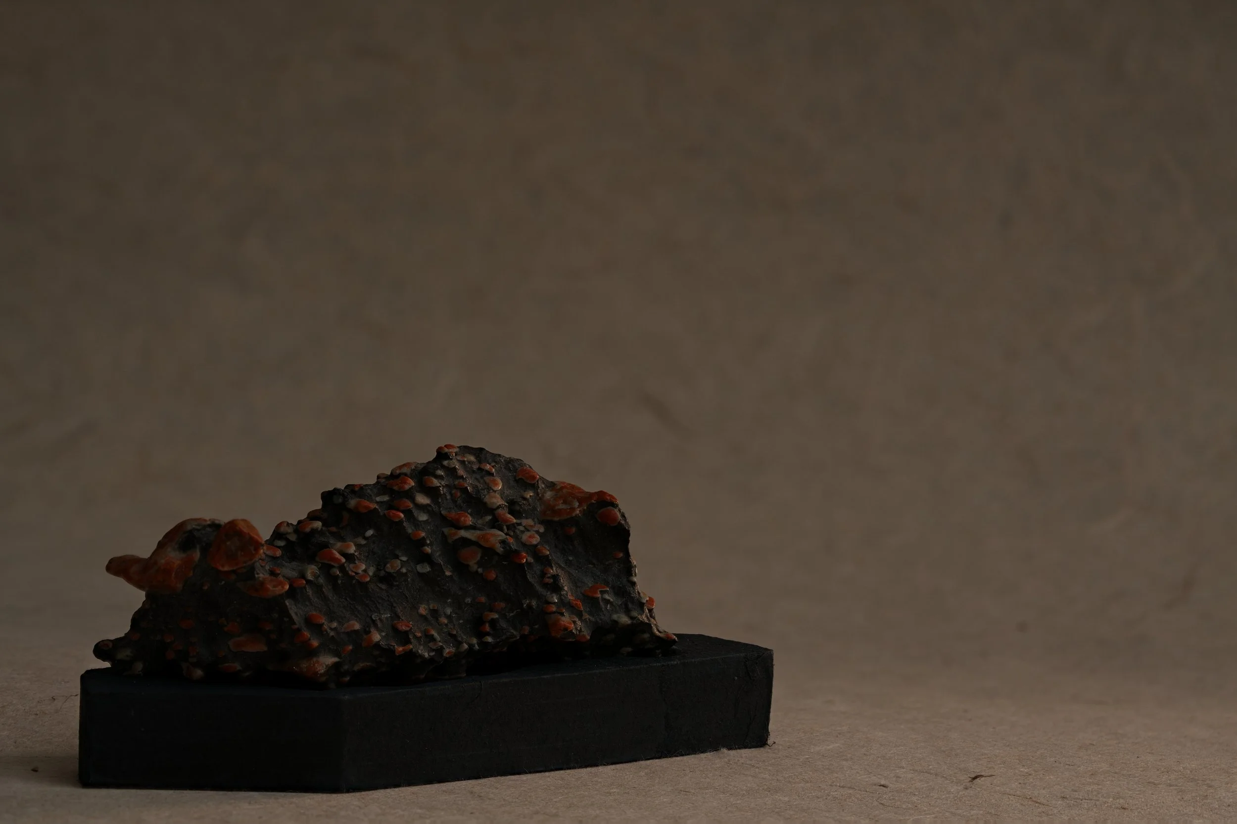 A rock or mineral specimen with black and red coloration and small red crystals, displayed on a black stand against a plain background.