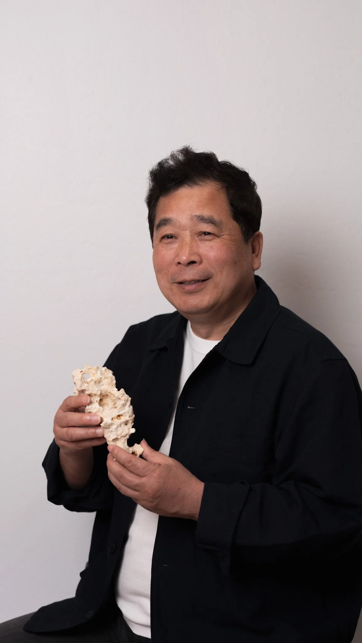 A man with dark hair, wearing a black jacket over a white shirt, sitting against a plain white wall, holding a large piece of coral.