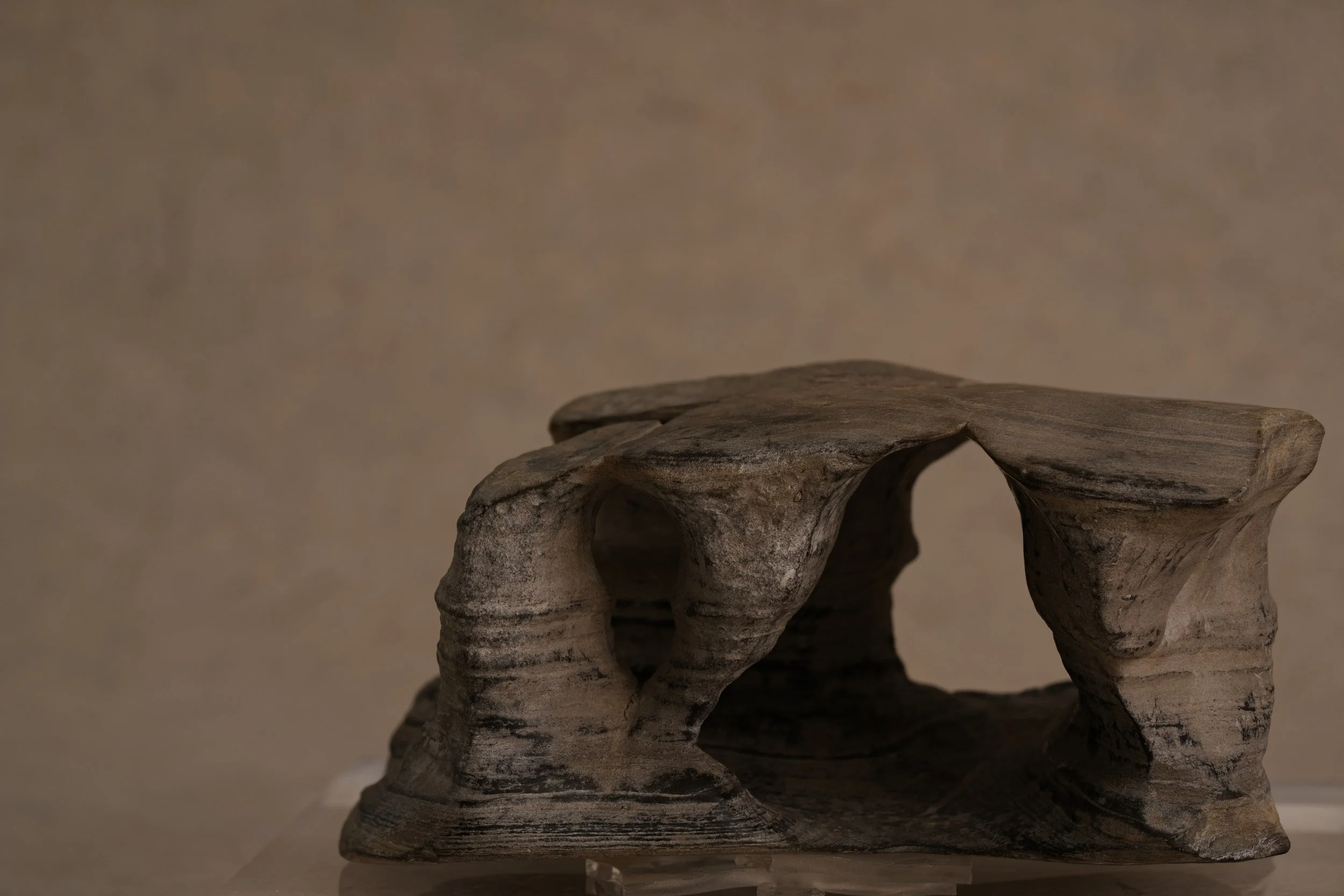 Ancient stone sculpture of a skull with hollow eye sockets, resting on a carved wooden base against a plain beige background.