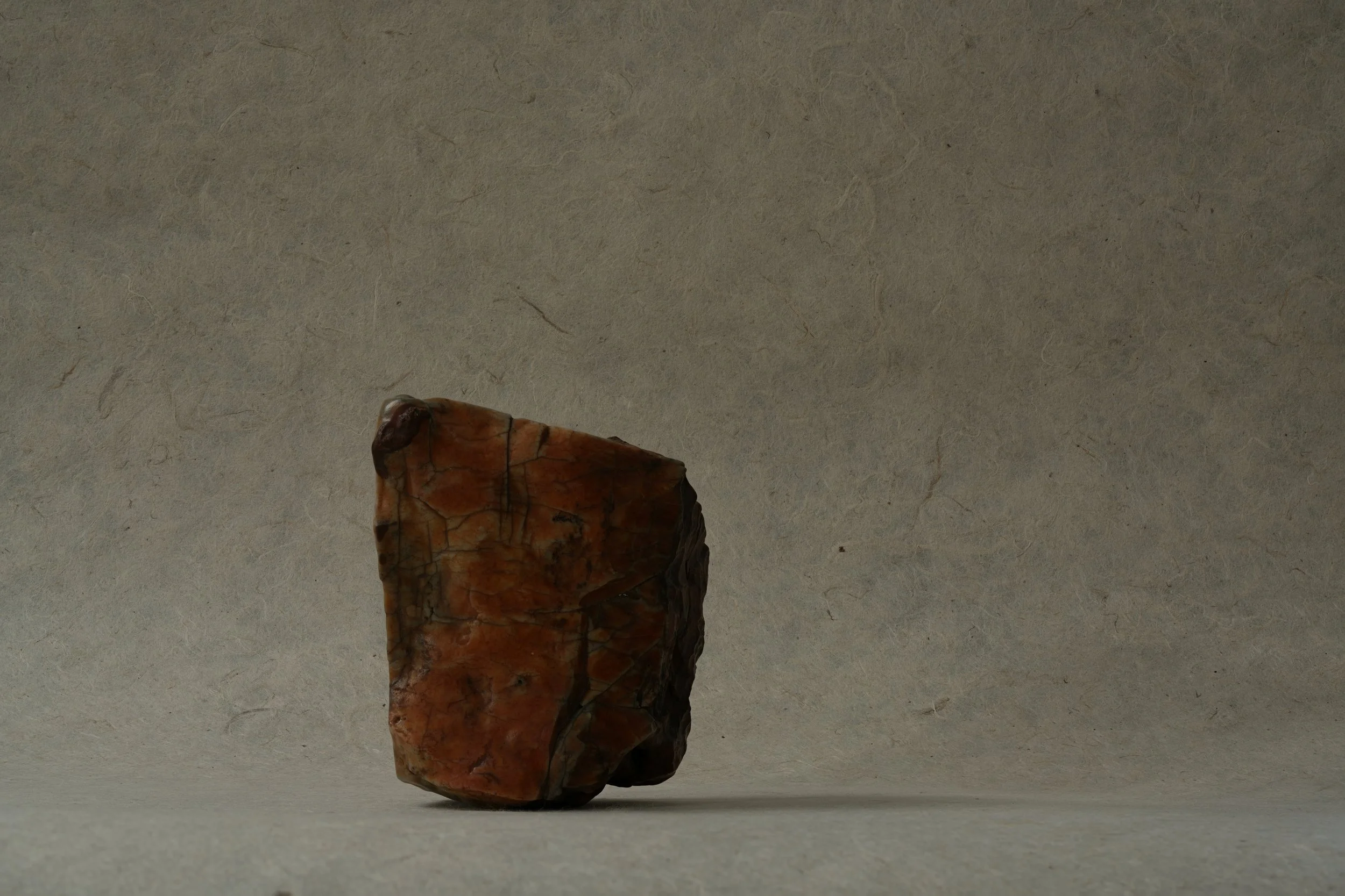 Close-up of a single brownish-red rock with a textured surface placed on a plain, light-colored surface, against a neutral background.