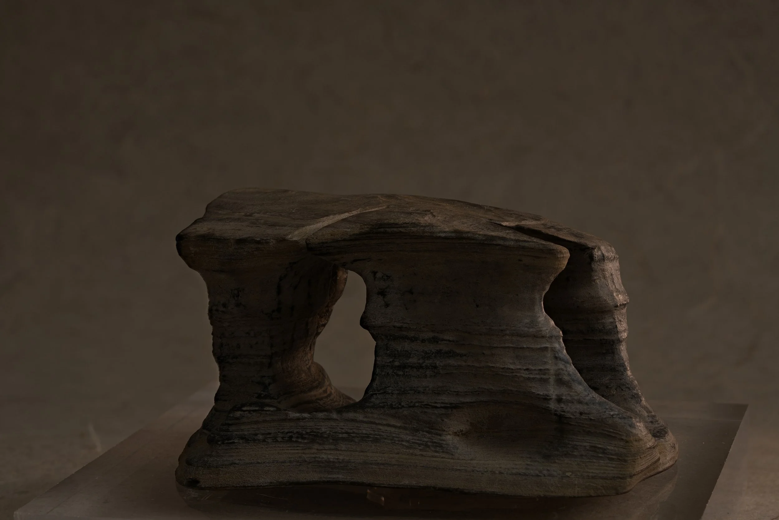 A layered, abstract sculpture made of dark, textured material resembling sedimentary rock, set against a plain background.