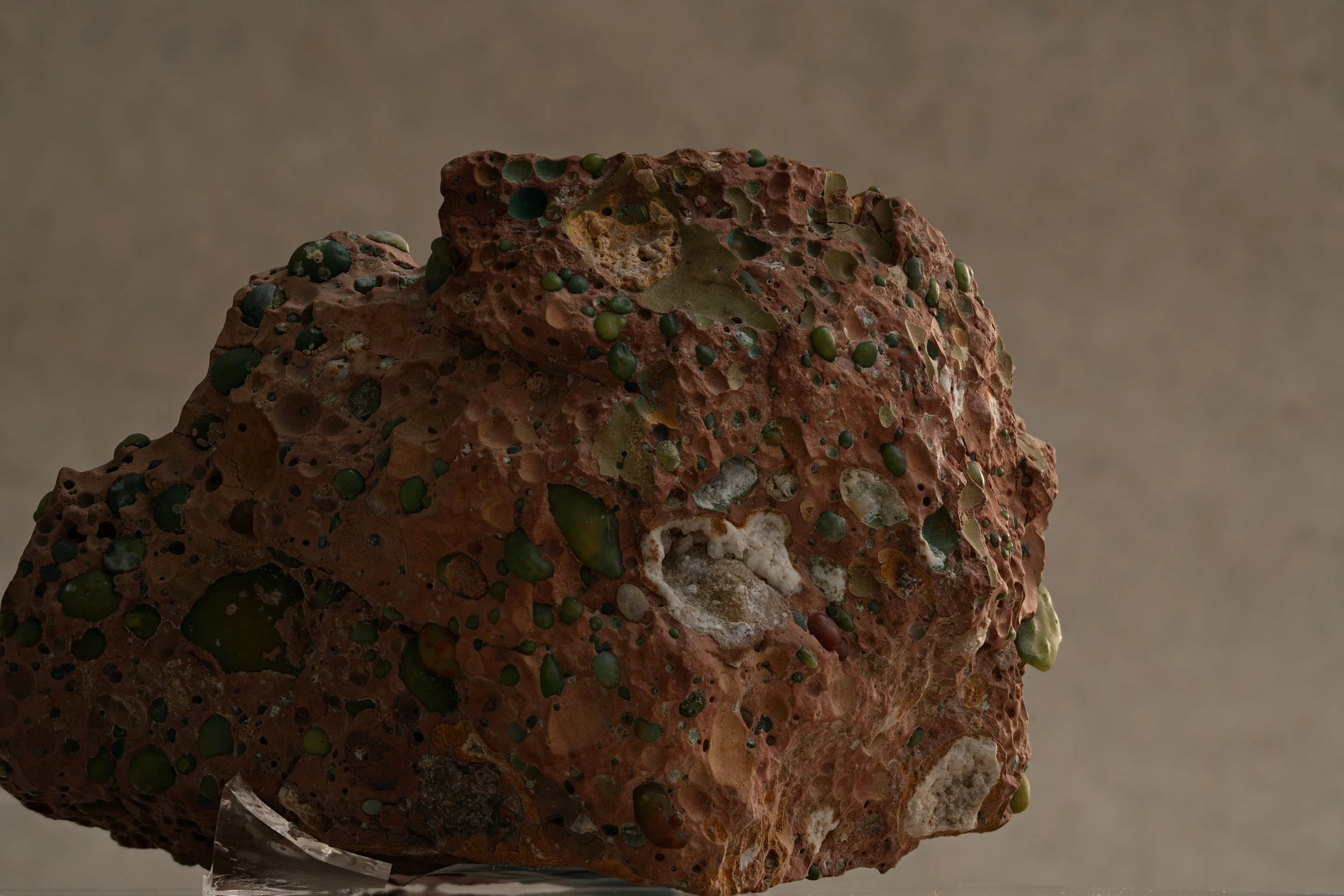 A close-up image of a rough, porous rock with green and dark-colored mineral deposits on its surface.