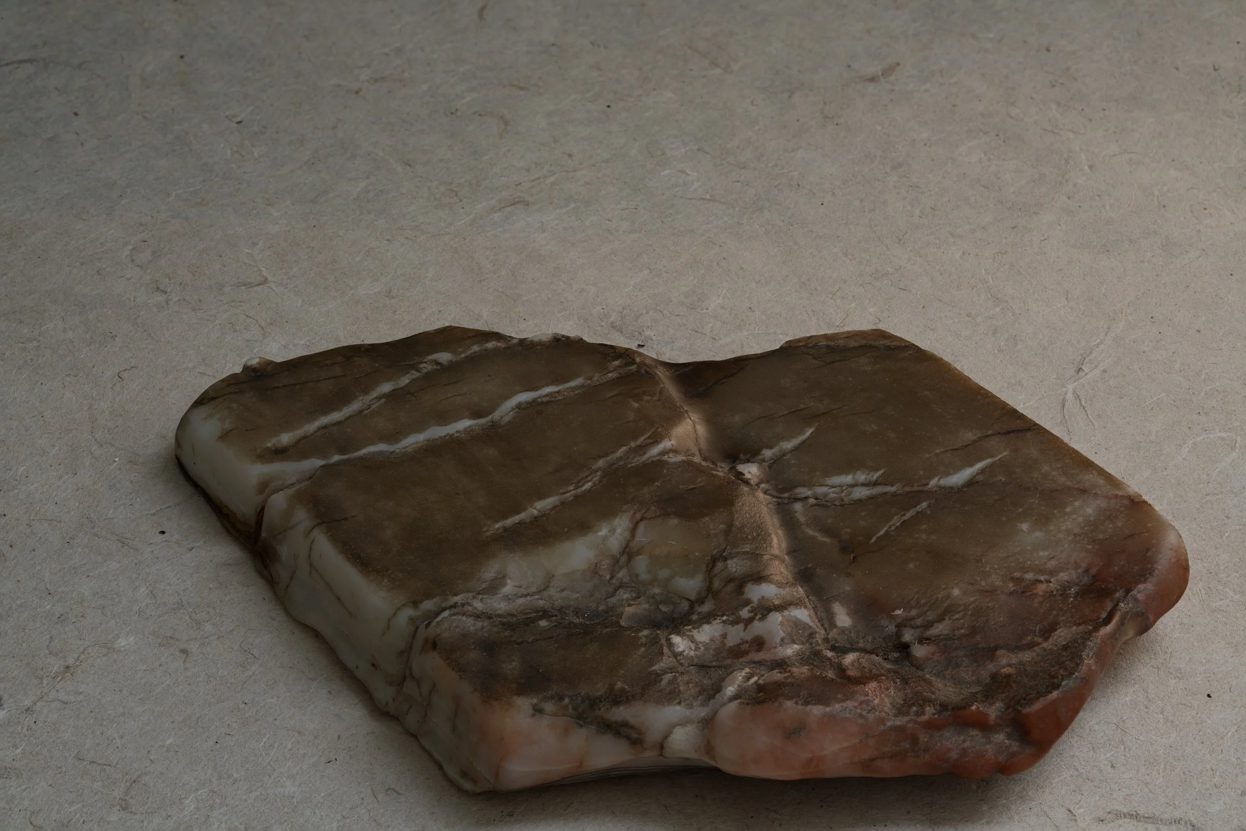 A rectangular block of soap with various brown and white marbling patterns, resting on a textured, light-colored surface.