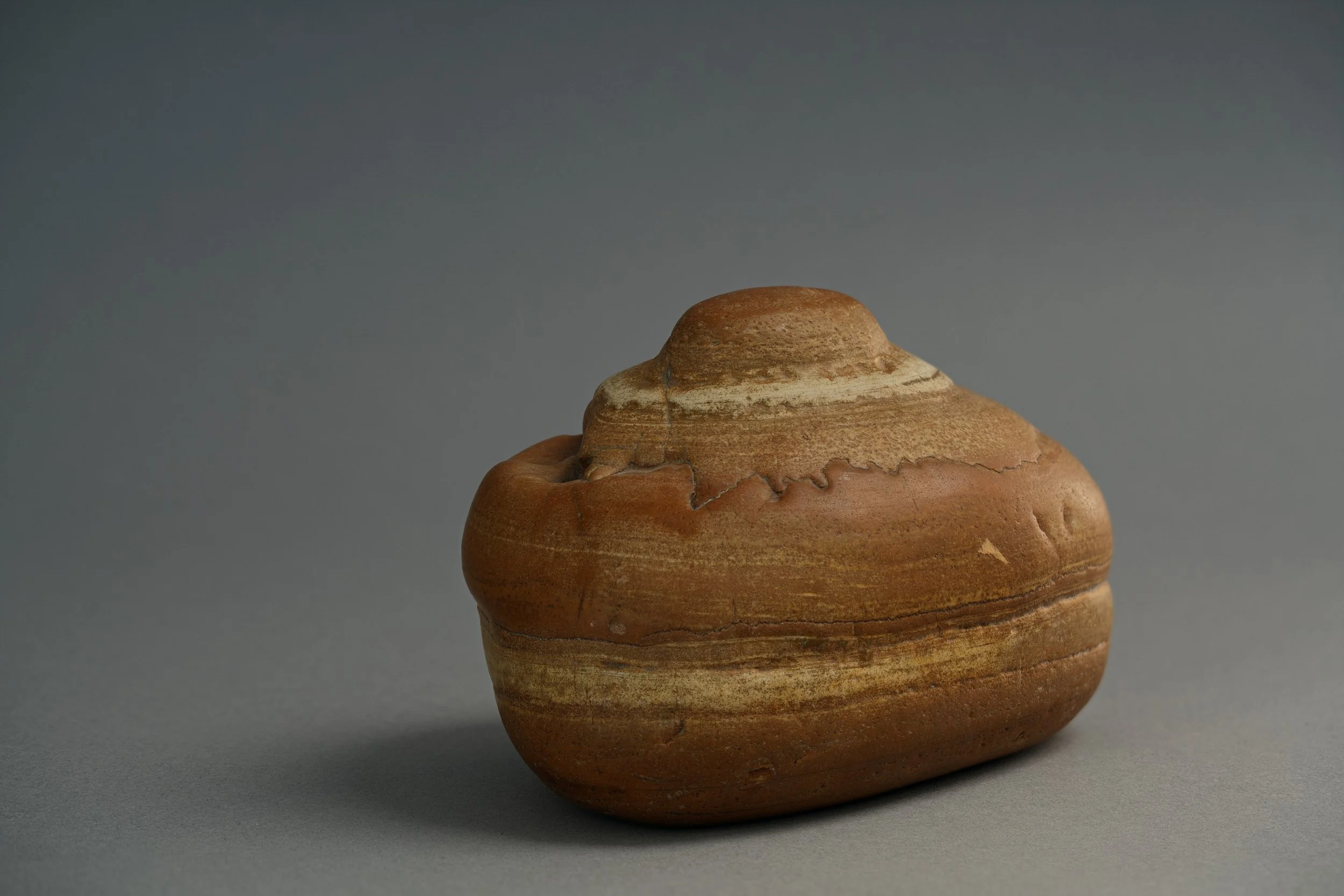 Close-up of a brown, rounded clay pottery vessel with horizontal striations and a small narrow opening on top, placed on a gray background.