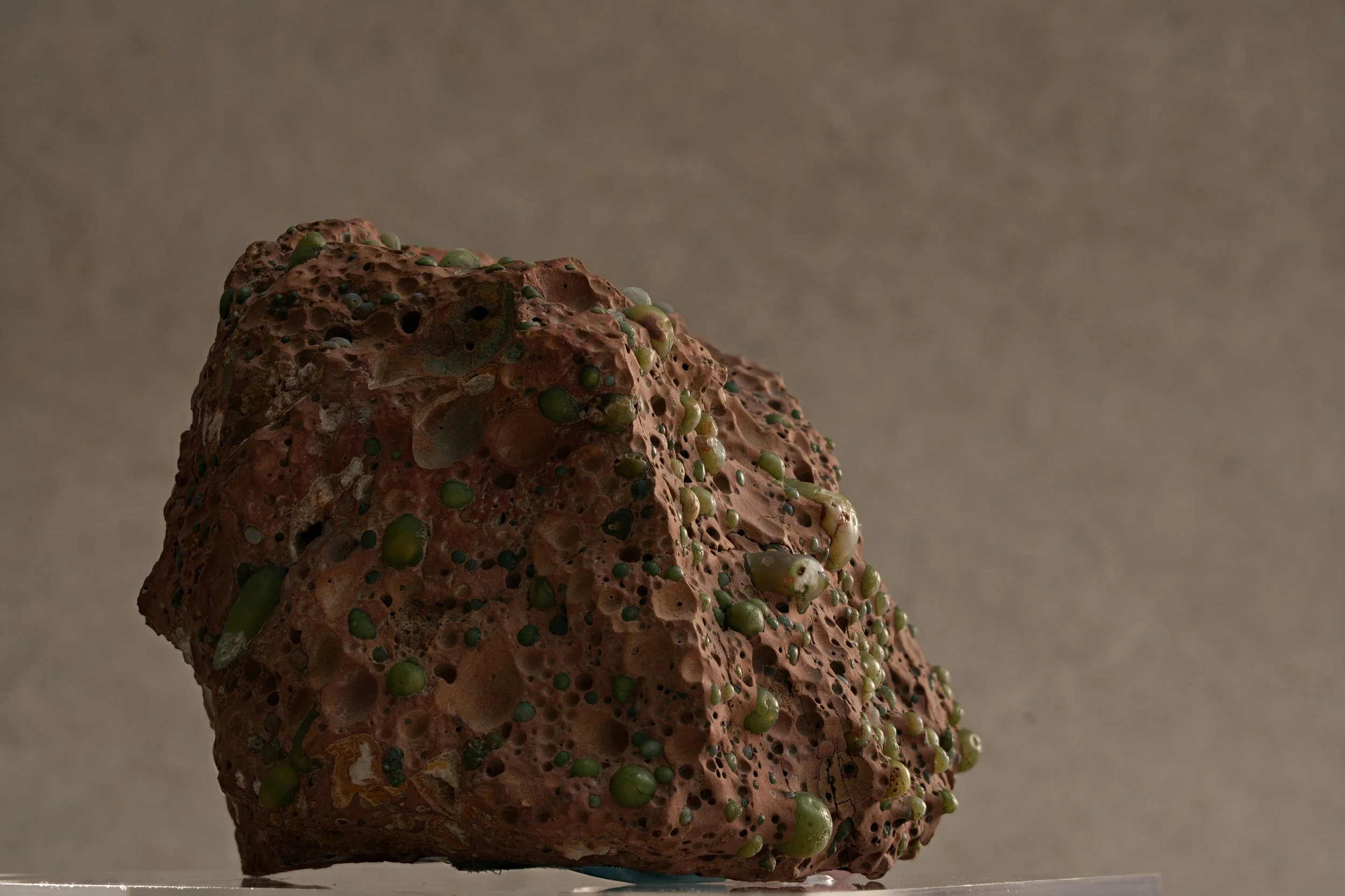 Close-up of a textured, reddish-brown volcanic rock with small greenish mineral deposits on its surface.