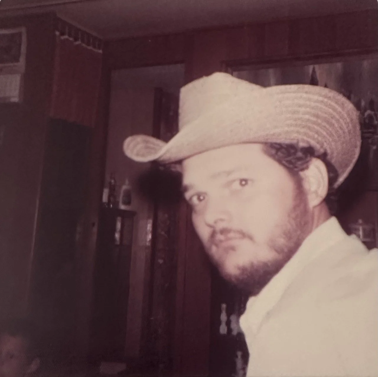 A man with a beard wearing a large straw cowboy hat and a light-colored shirt, looking at the camera, indoors with dark wood paneling and shelves in the background.