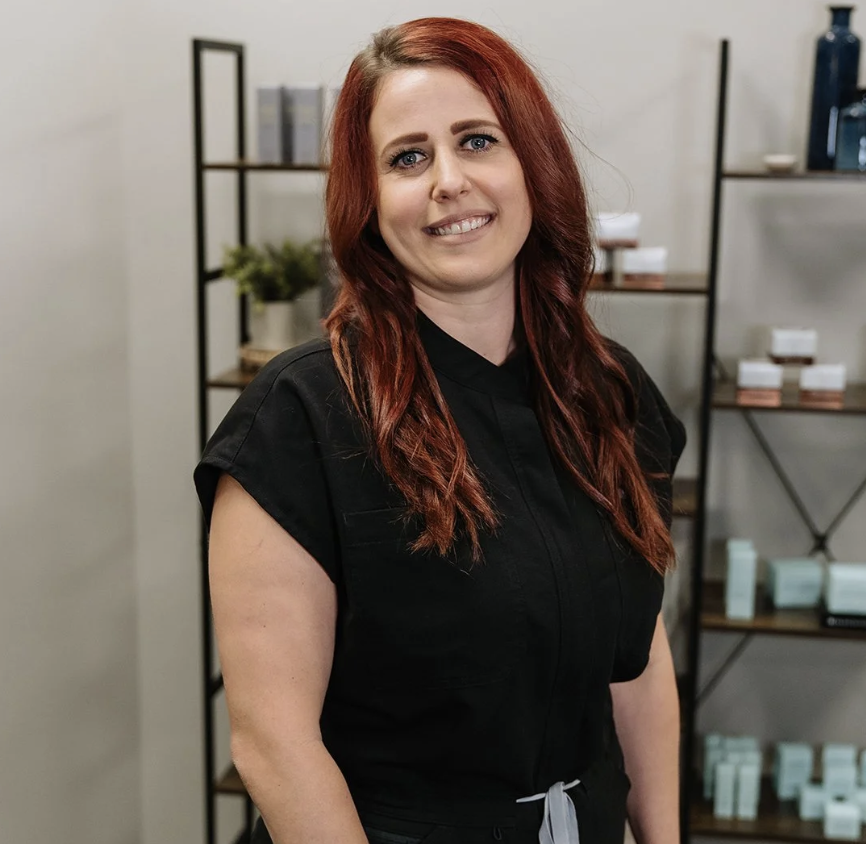 A woman with long red hair, blue eyes, and a black uniform, standing in a room with shelves holding various skincare or cosmetic products.