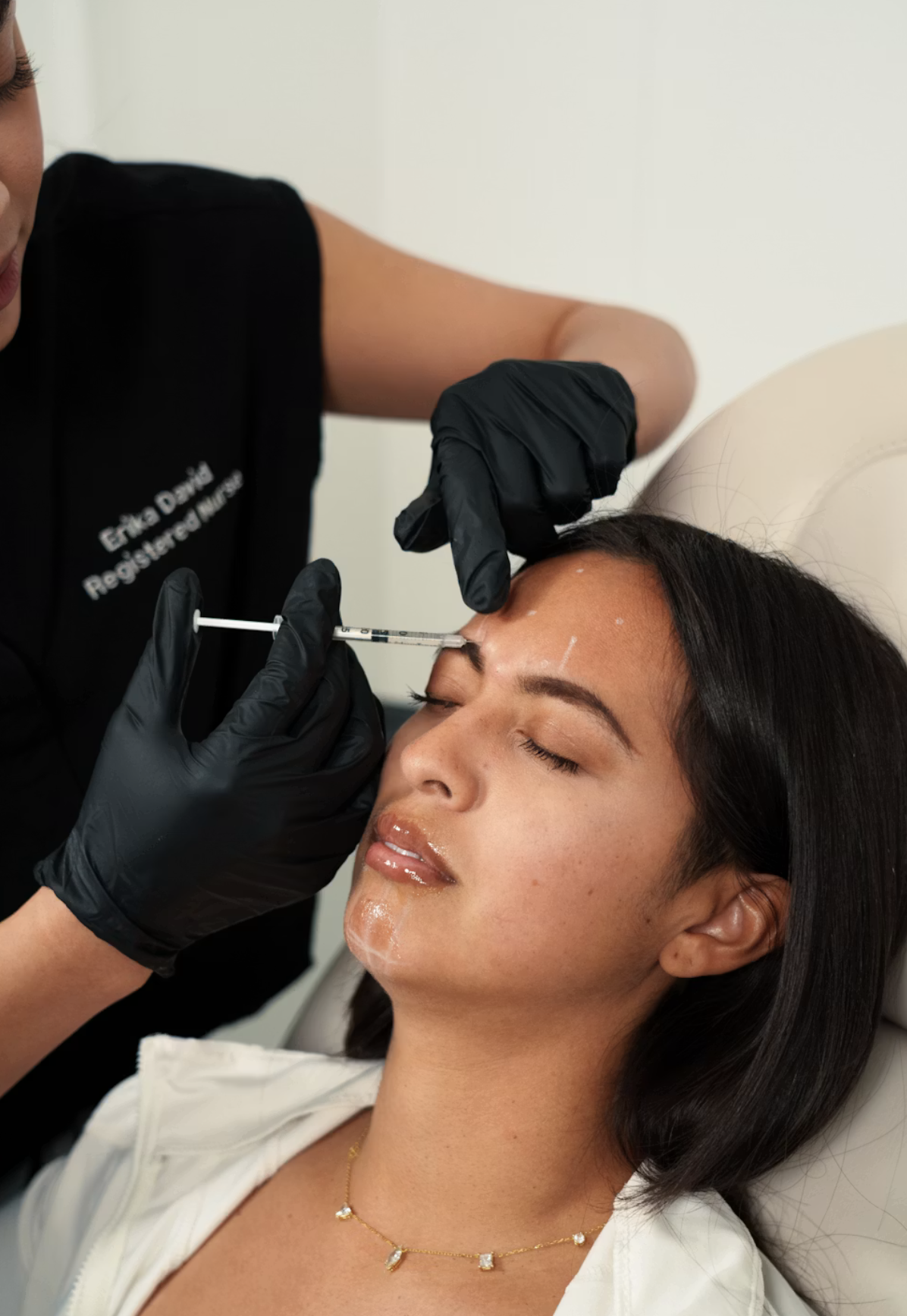 A woman receiving a cosmetic injection on her forehead by a medical professional wearing black gloves.