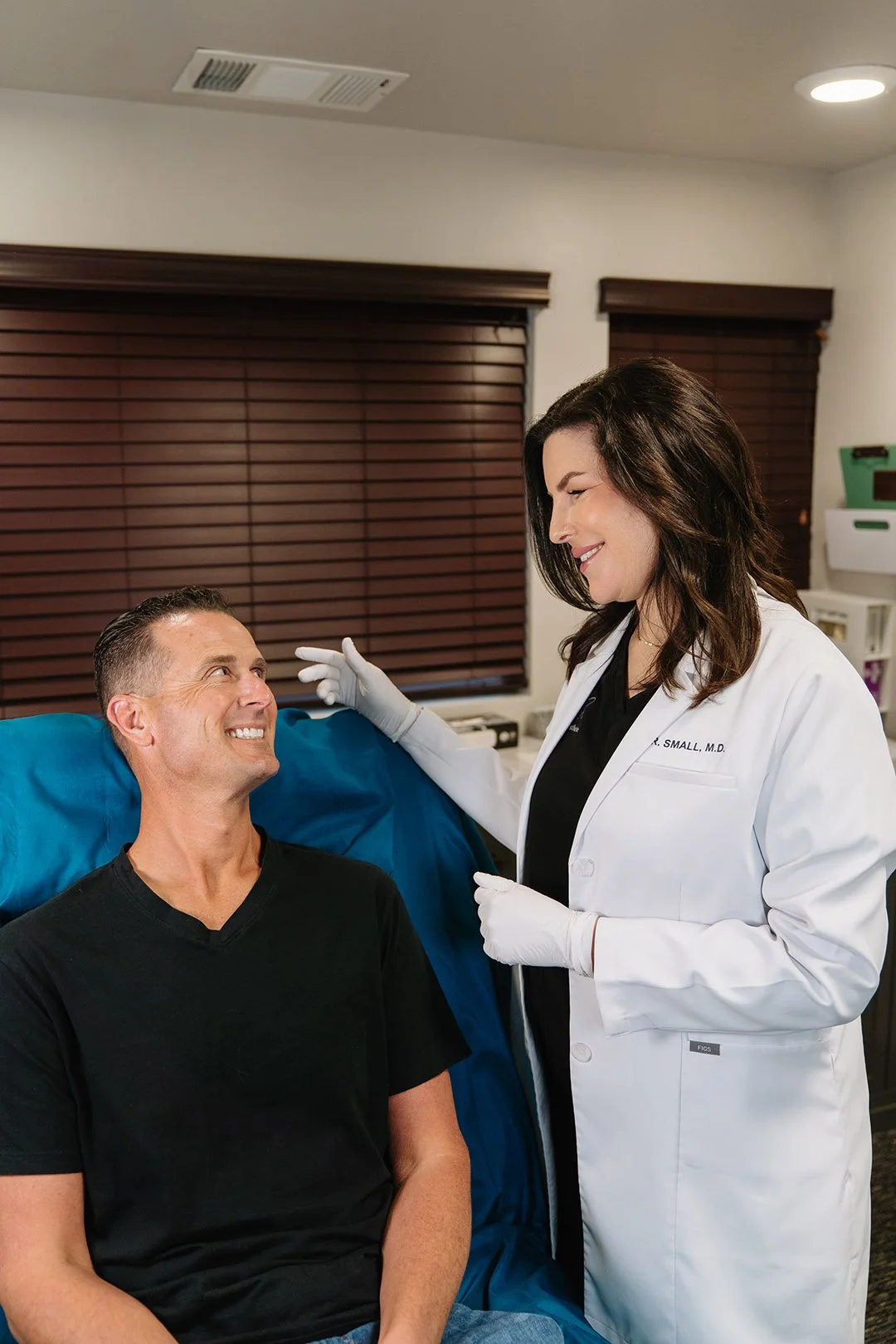 A smiling male patient in a black T-shirt sitting on a medical examination table, talking to a female doctor in a white coat and gloves in a medical office with wooden blinds.