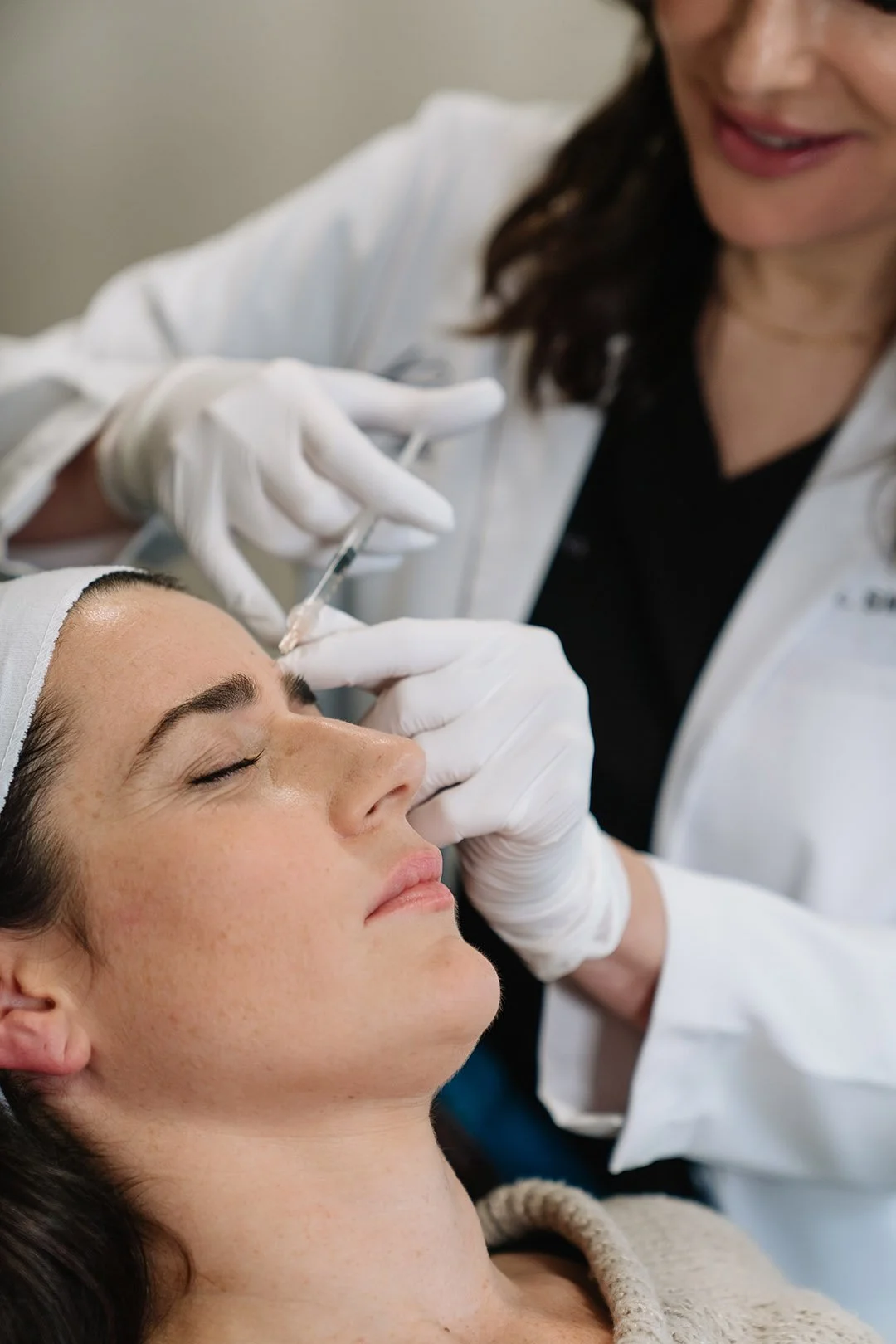 A woman receiving a cosmetic injection in her forehead from a healthcare professional wearing gloves in a clinical setting.