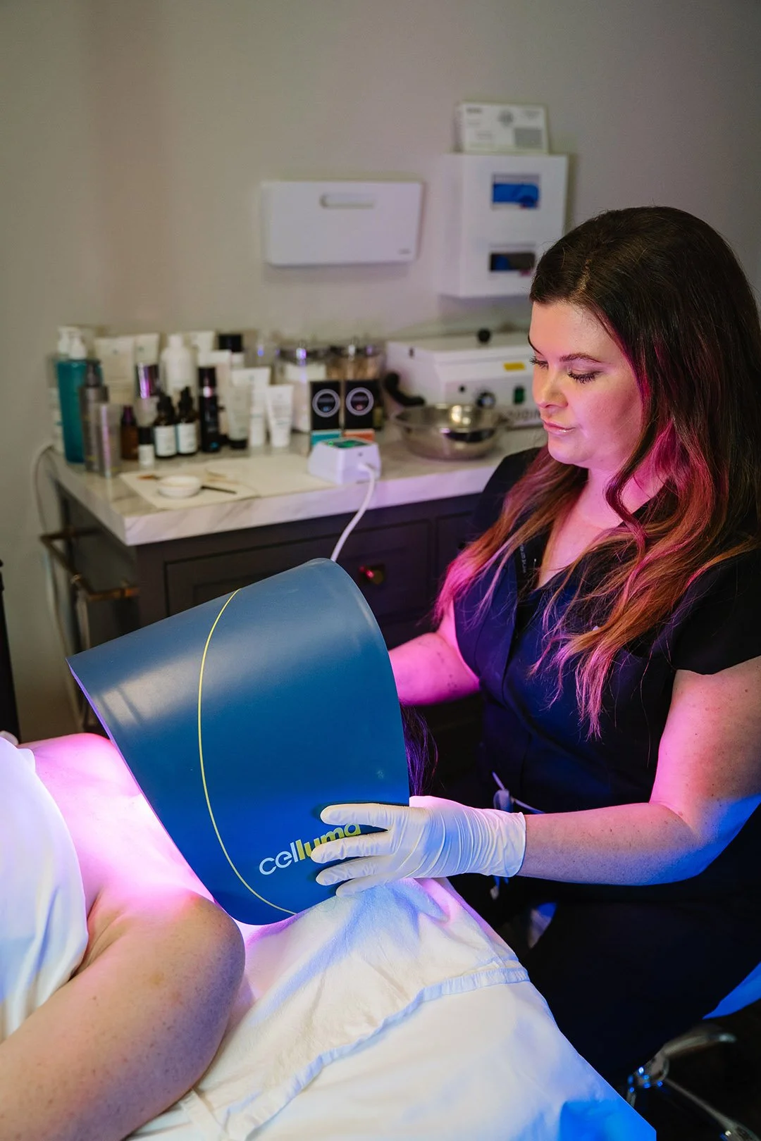 A woman in black uniform wearing gloves performing a skincare treatment with a handheld LED light device on a patient lying on a bed, in a clinic or spa setting with skincare products and equipment in the background.