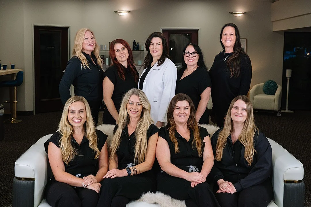 Group of nine women in professional attire posing together in a room, some standing and some sitting.