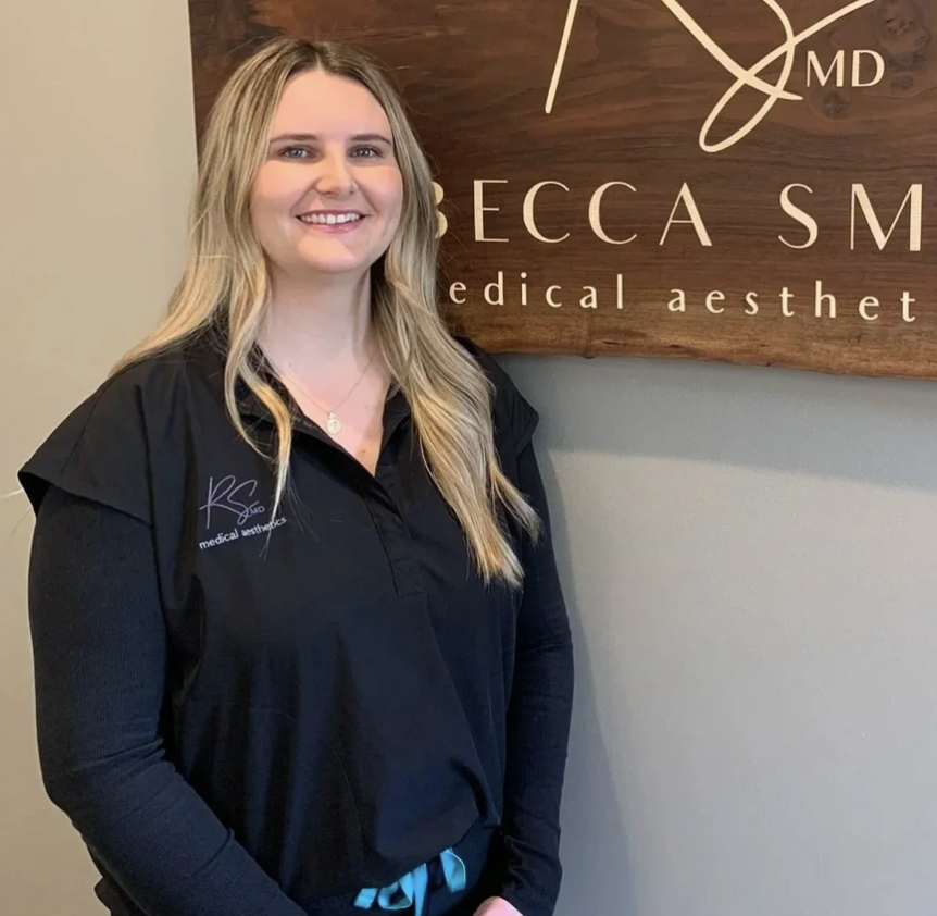 Young woman with long blond hair smiling, standing in front of a wooden sign that reads 'Becca SM medical aesthetic' and wearing a black uniform with her name and logo embroidered on it.