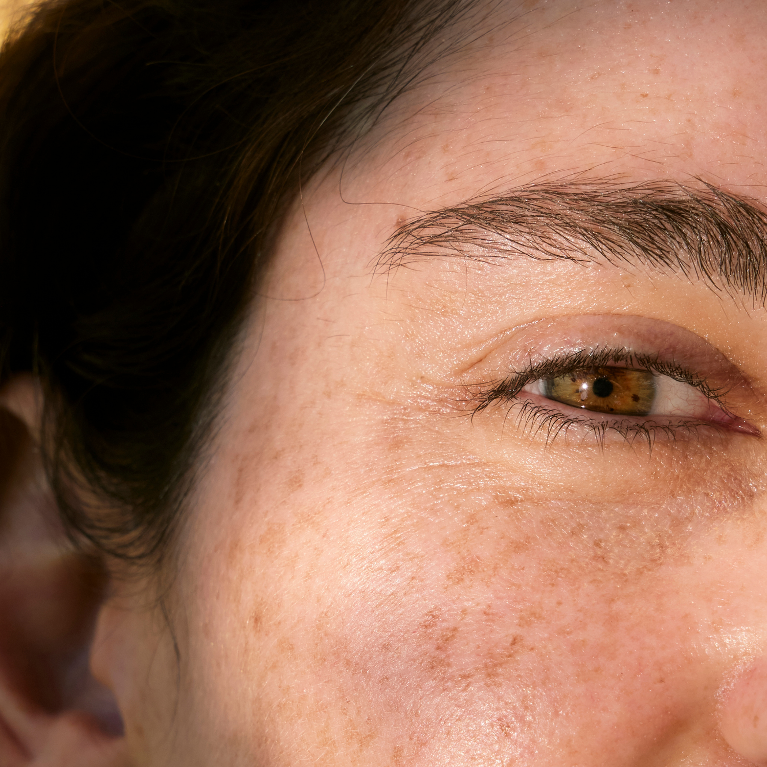 Close-up of a woman's face showing her brown eye, eyebrow, and surrounding skin with freckles and fine details.