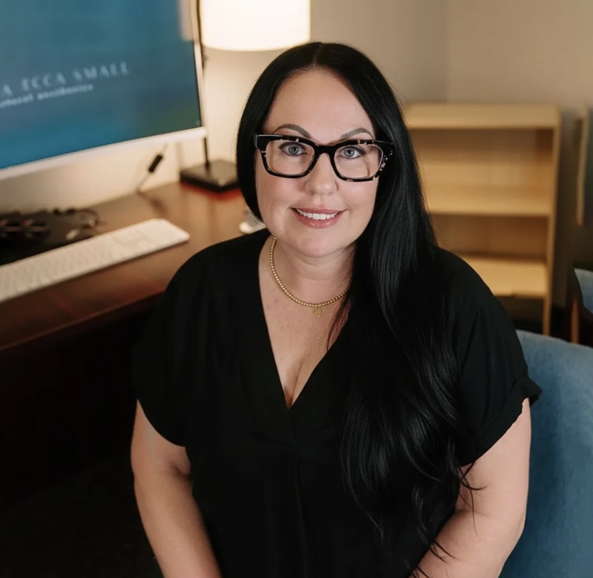 A woman with long black hair, glasses, and a gold necklace smiling while sitting in a room with a desk, computer monitor, lamp, and bookshelf in the background.