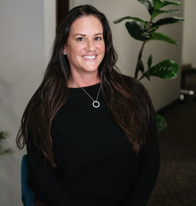 Smiling woman with long dark hair in a black top, standing indoors with a plant in the background.