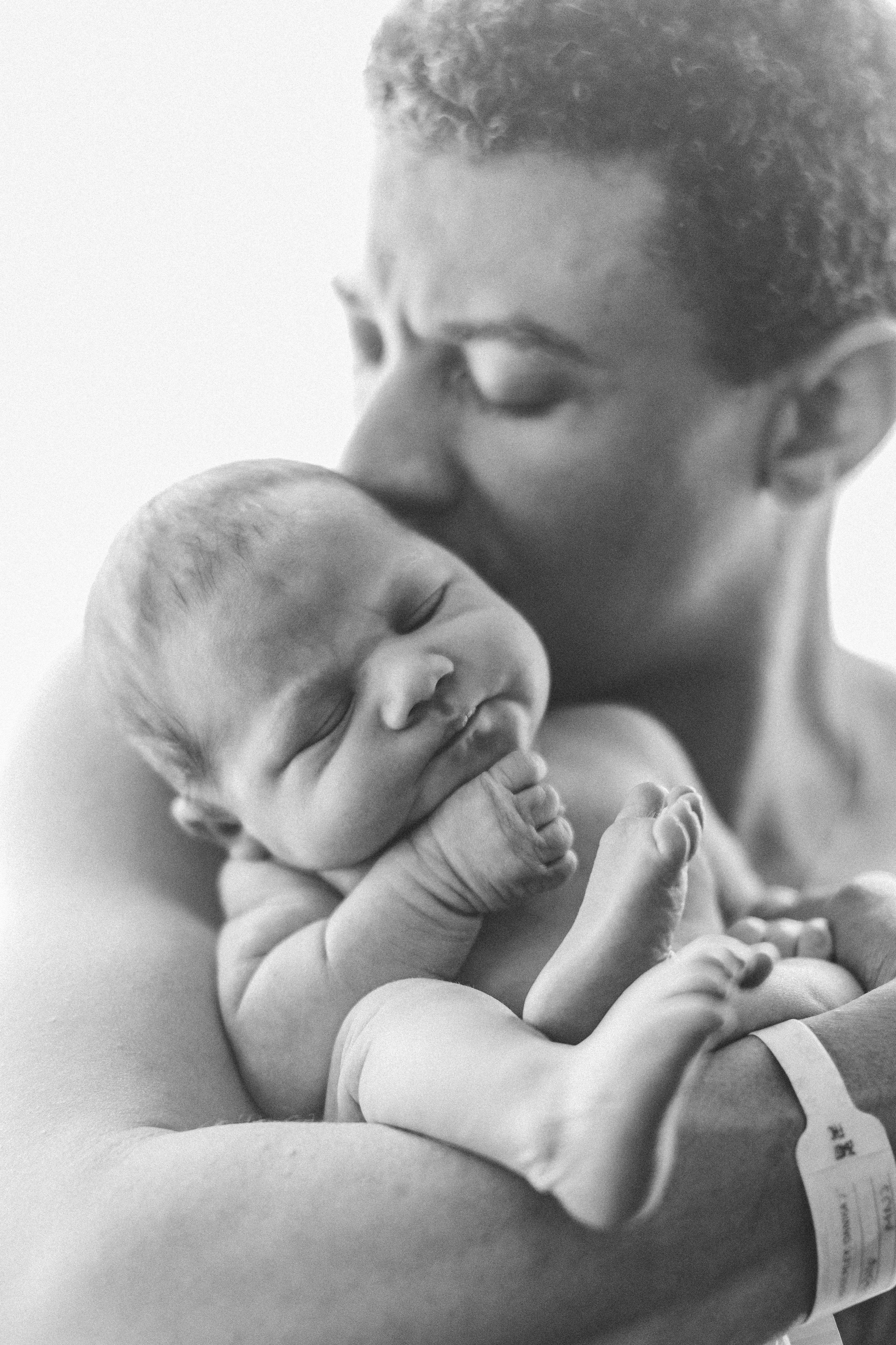 A newborn baby sleeping peacefully on an adult's arms, with a woman kissing and holding the baby, in black and white.