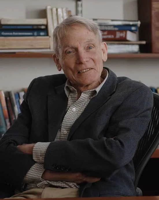 An elderly man with gray hair, wearing a checkered shirt and blazer, sitting in an office with bookshelves in the background.