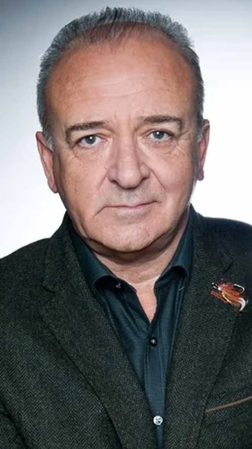 Headshot of a middle-aged man with short gray hair, wearing a dark blazer and a dark shirt, against a plain background.