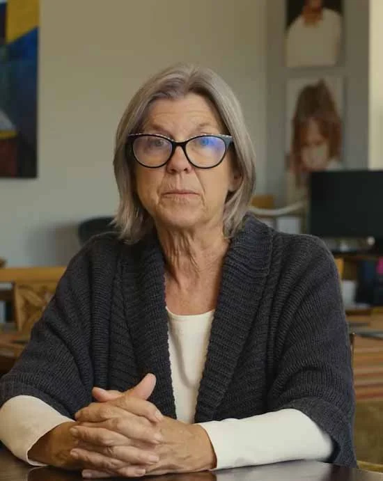 An elderly woman with gray hair and glasses sitting at a desk in an office or home setting.