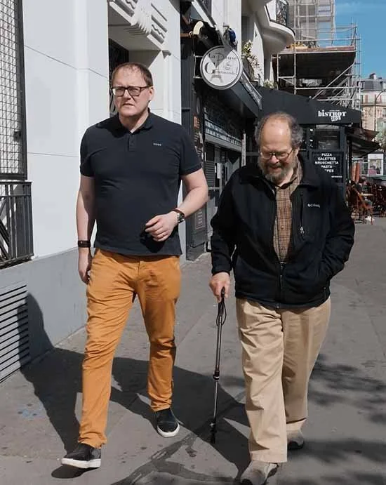 Two men walking on a city sidewalk, one in black shirt and orange pants, the other with a cane in a black jacket and beige pants, with storefronts and buildings in the background.