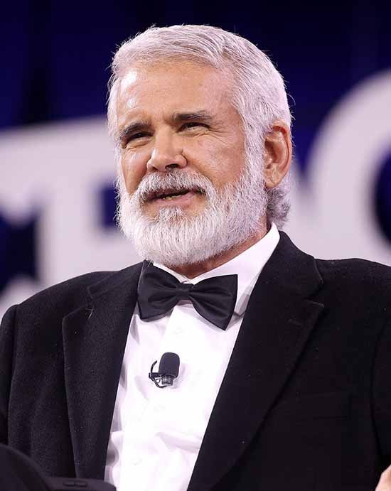 A man with gray hair and a beard wearing a tuxedo and bow tie, speaking at a formal event.