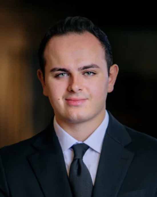 A young man in a dark suit, white shirt, and black tie smiling confidently.