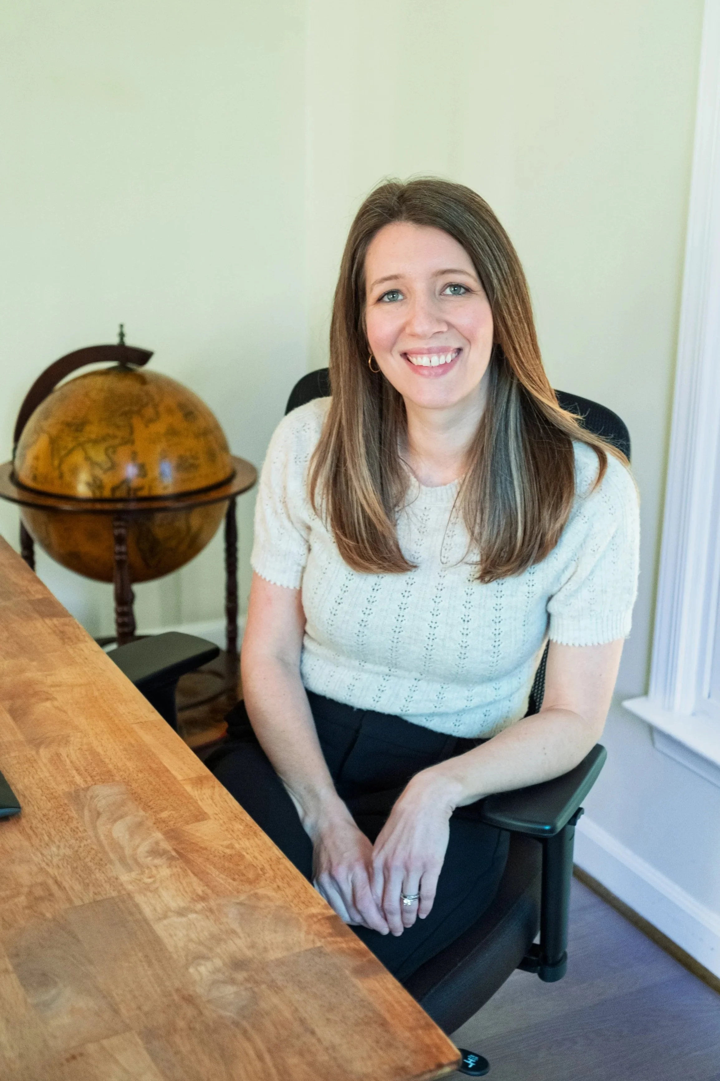 A woman with brown hair smiling, sitting at a wooden desk with a globe beside her in a room with white walls.