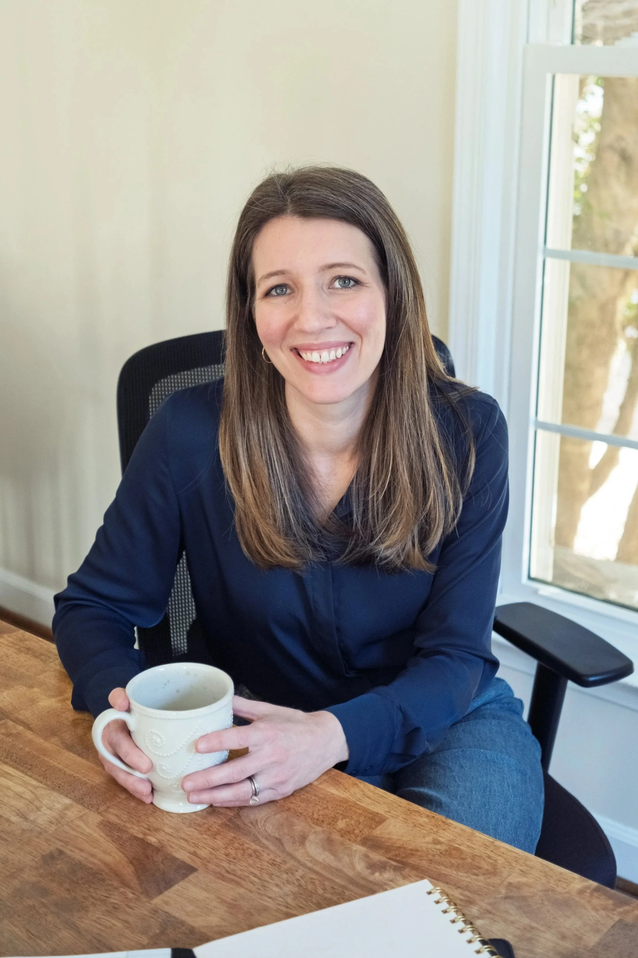 A woman with long brown hair, wearing a dark blue blouse, sitting at a wooden table with a ceramic coffee mug in her hands, smiling, near a window with sunlight and a view of trees outside.