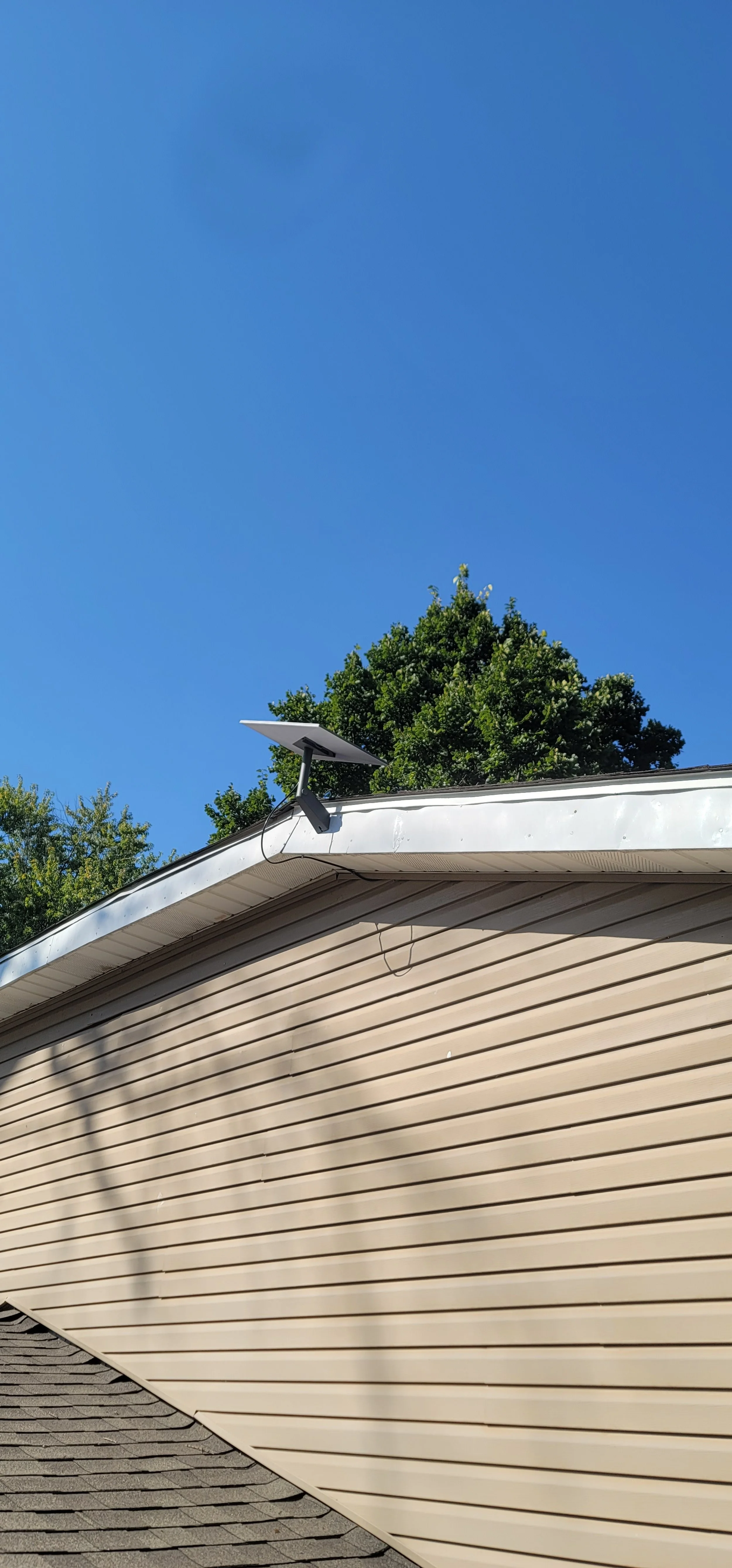 Photo of the side of a house with beige vinyl siding, a gray shingled roof, a white gutter, and a solar panel mounted on the roof. In the background, clear blue sky and green trees are visible.