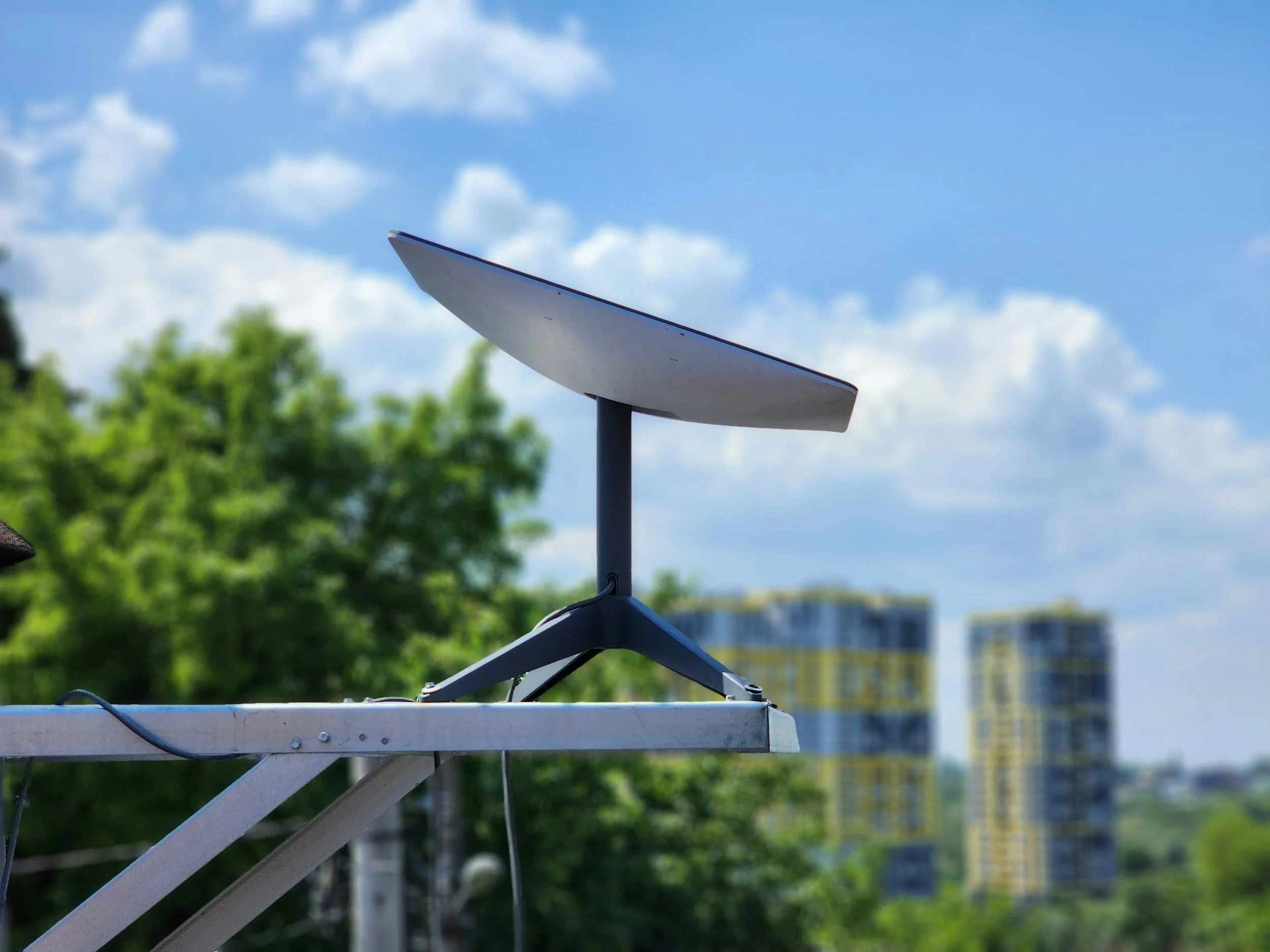 Satellite dish mounted on a rooftop against a partly cloudy sky, with a background of green trees and distant buildings.