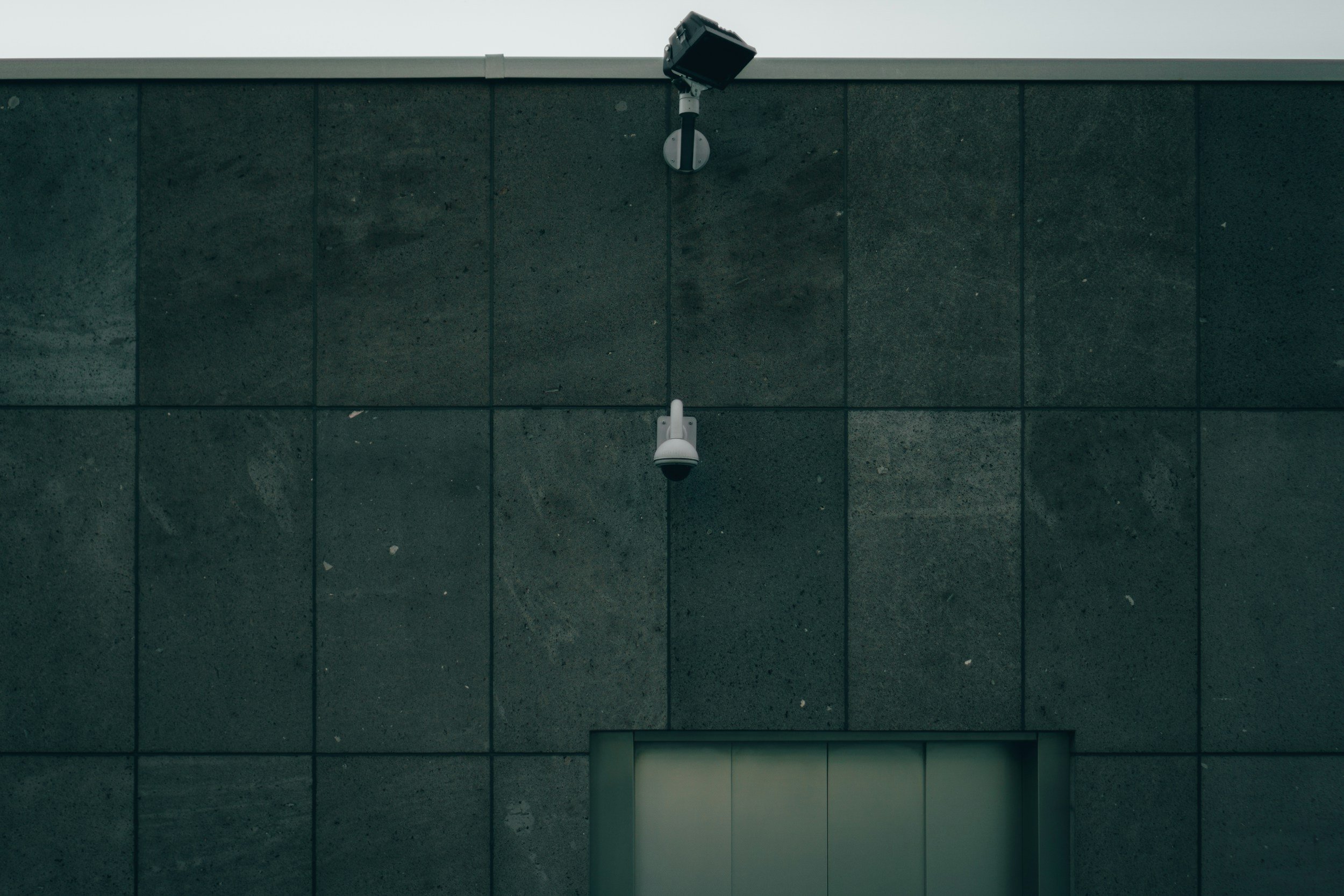 Close-up of a modern building wall with a security camera and a small device mounted above a rectangular opening.