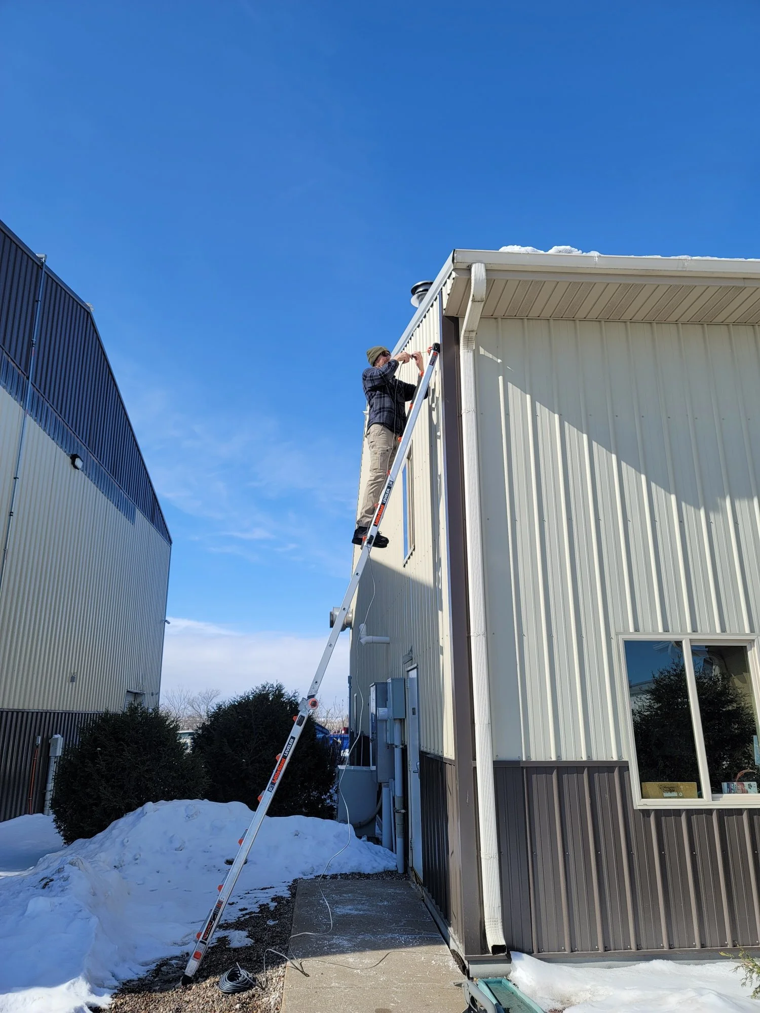A person on a ladder working on the exterior wall of a building under a clear blue sky, with snow on the ground.