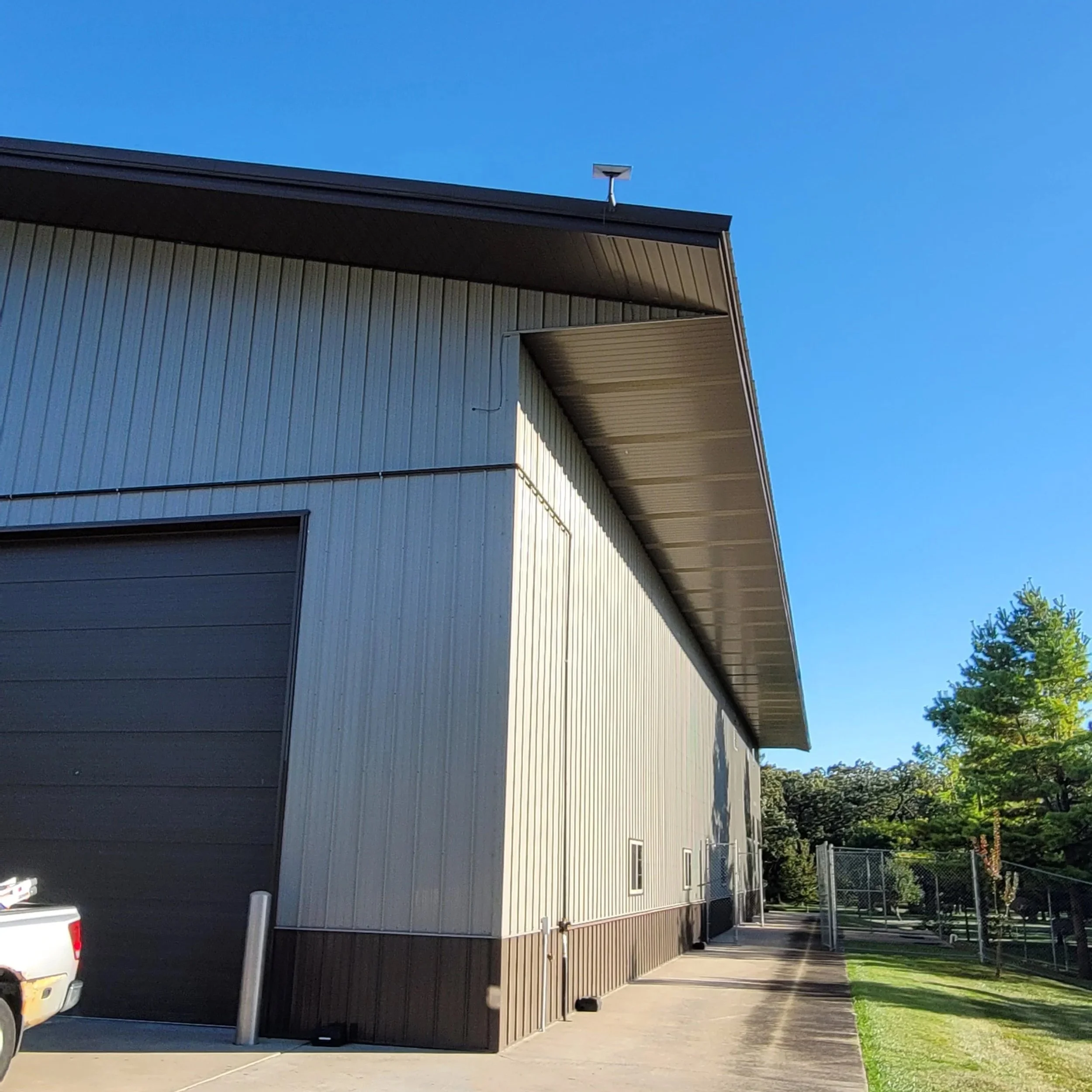 Side view of a building with beige metal siding, a large closed garage door, and a concrete pathway. There is a white truck partially visible on the left, a fence on the right, and trees behind the fence. The sky is clear and blue.