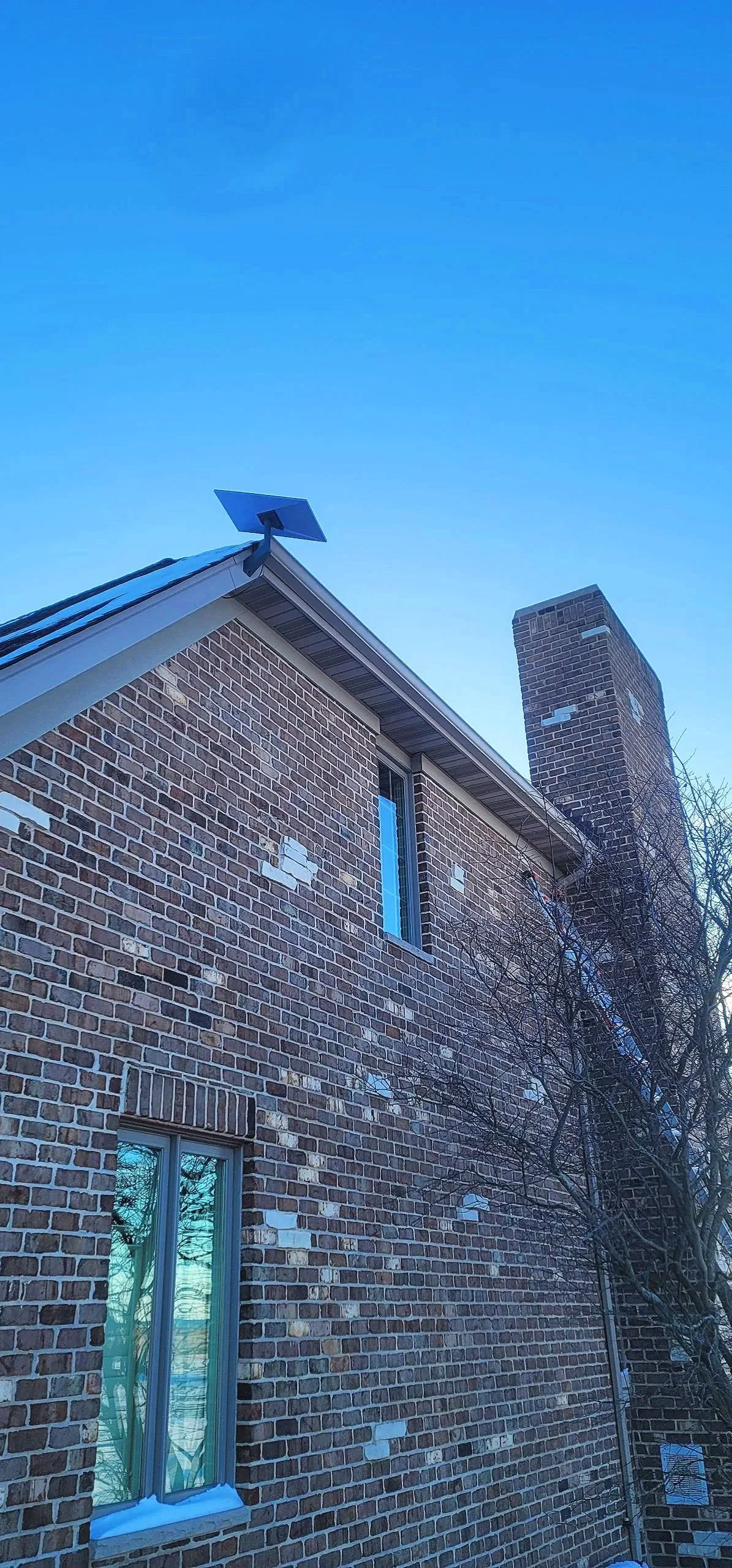 A brick house with a chimney, three windows, and solar panels on the roof, against a blue sky.