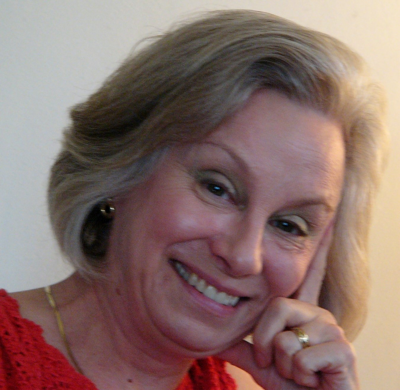 A smiling woman with blonde hair styled in waves, wearing a red top and gold jewelry, resting her face on her hand.