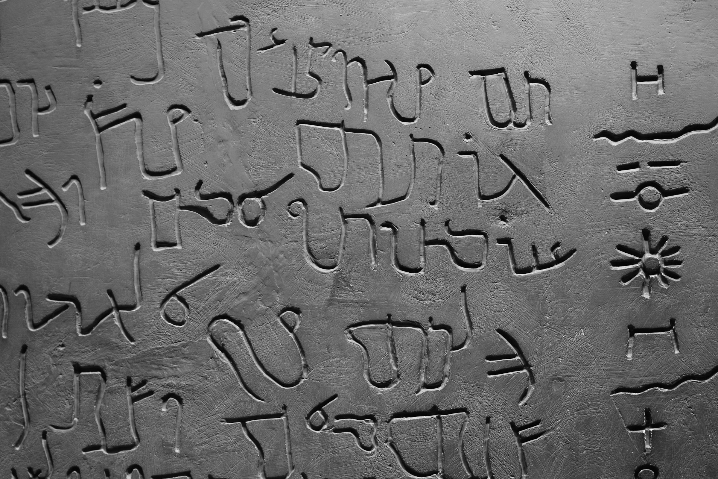 Close-up photograph of a stone or metal surface with inscriptions or carvings, featuring various symbols and characters, some resembling letters or scripts, with a textured background.