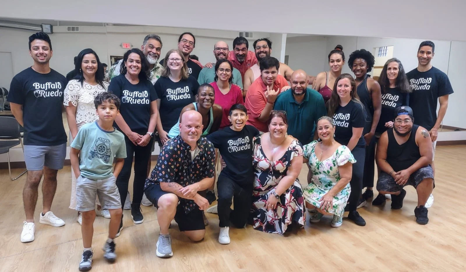 Group of people posing for a photo in a room with a wooden floor, some wearing black t-shirts that say 'Buffalo Rueda', others in casual clothing, smiling and looking at the camera.