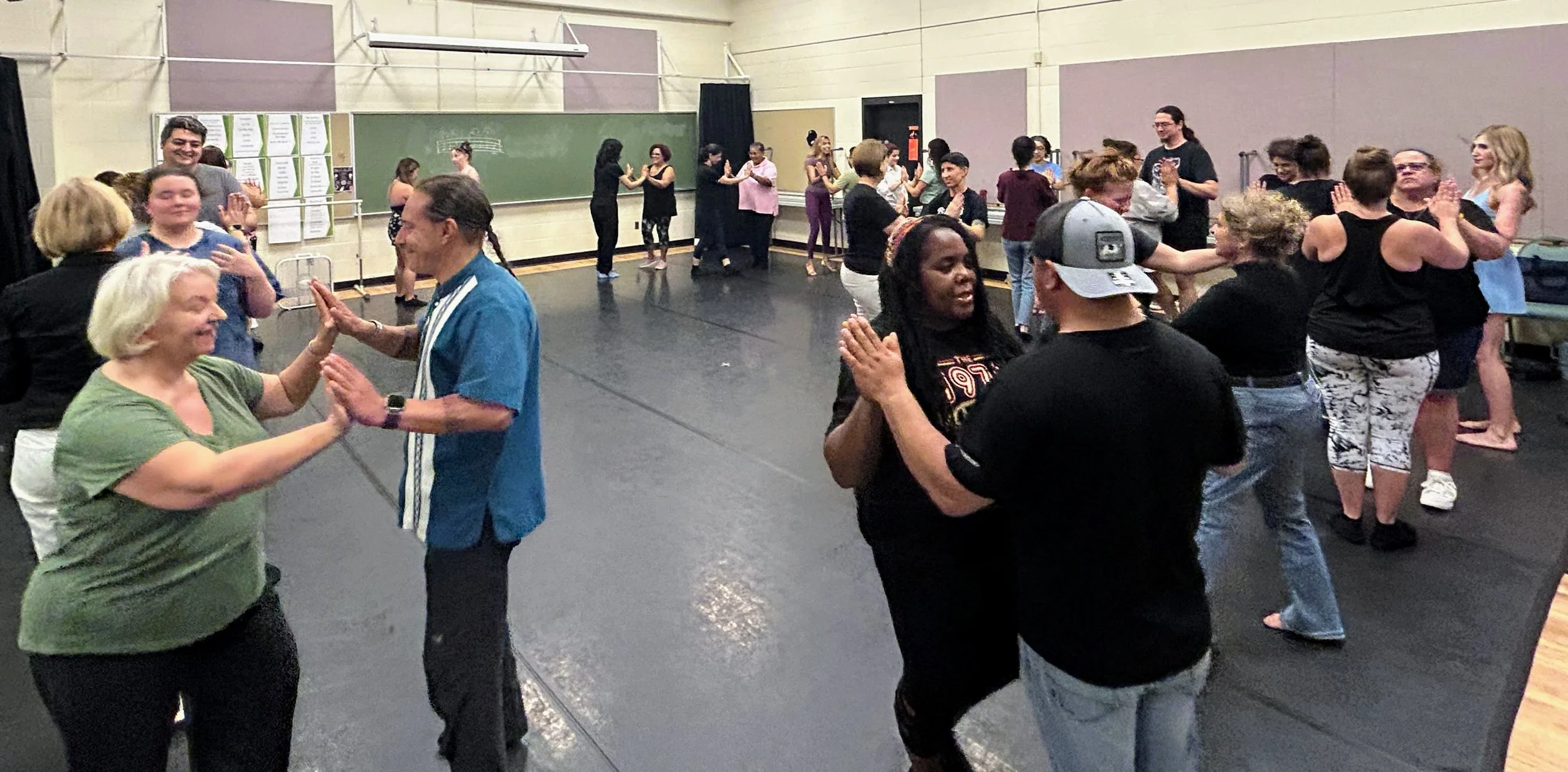People dancing and socializing in a dance class or social event in a gym or dance studio with black flooring, beige walls, and a chalkboard.
