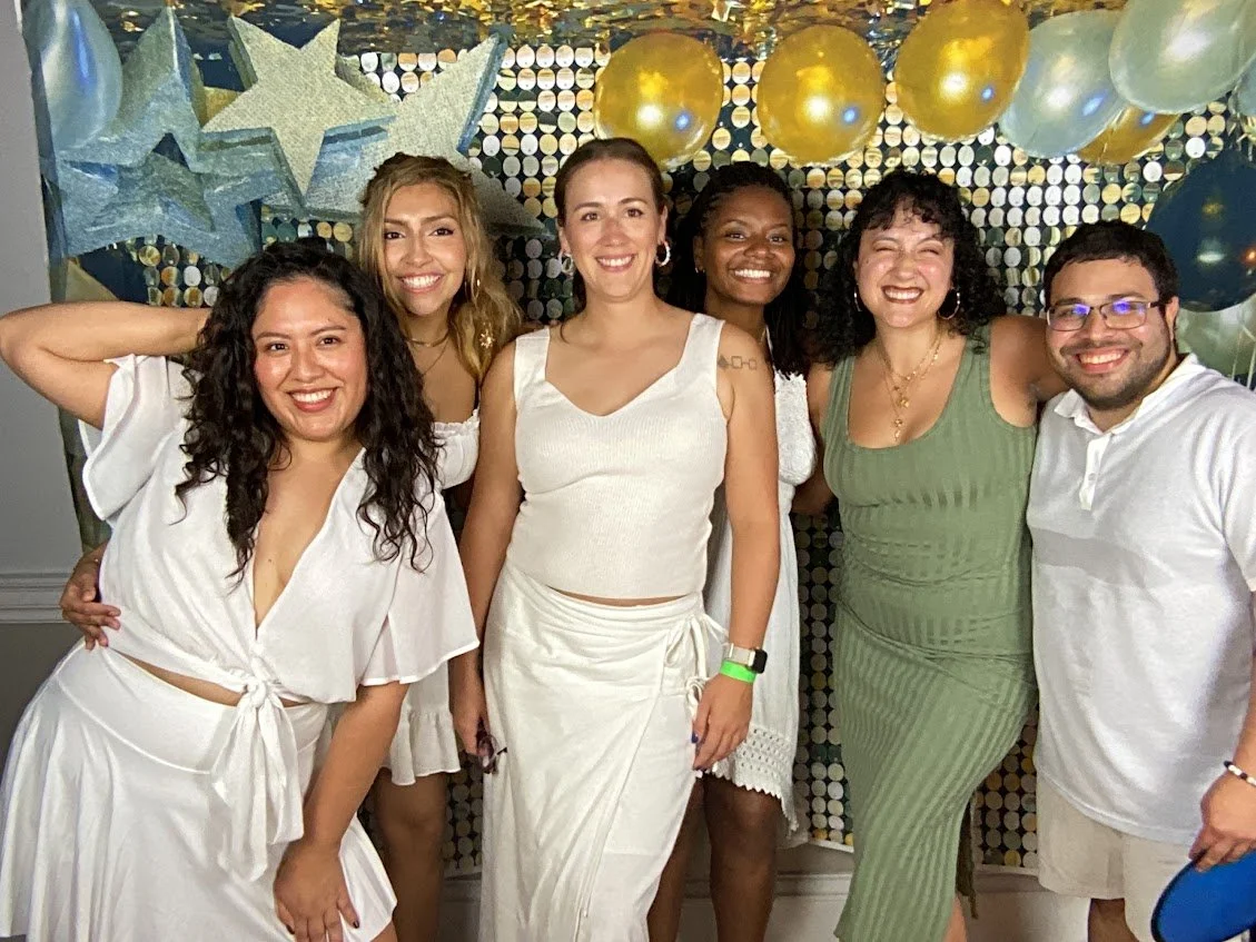 Group of six diverse women and one man posing together at a celebration with star and balloon decorations in the background