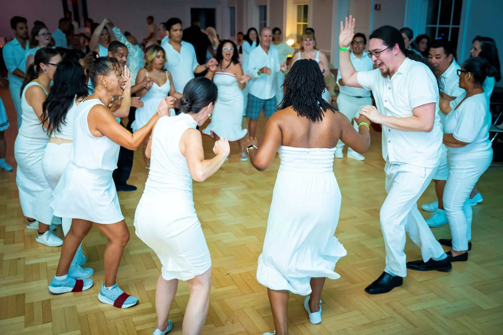 People dancing and celebrating at a party or event, all dressed in white, with some wearing casual and others in wedding or formal attire, in a well-lit indoor venue with wooden flooring.