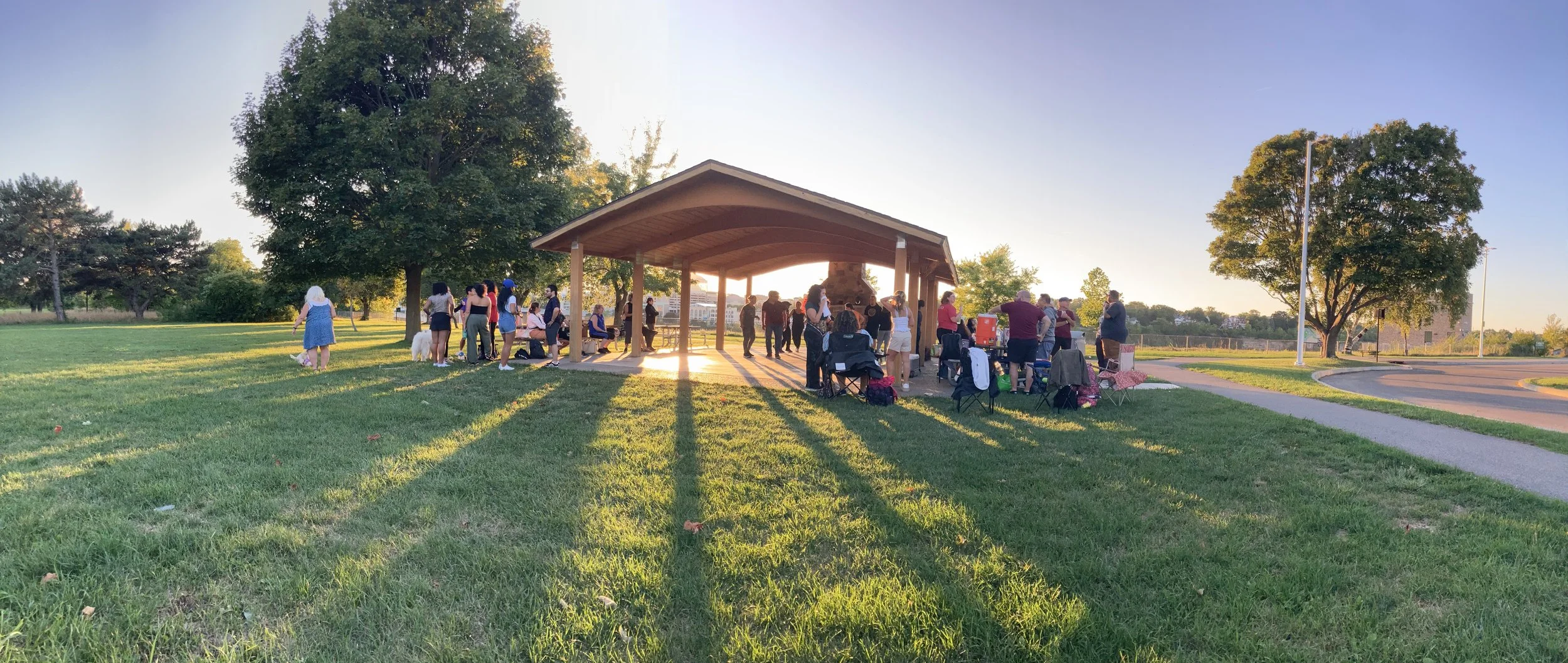 People gathered under a pavilion in a park during sunset, with chairs and trees in the background.